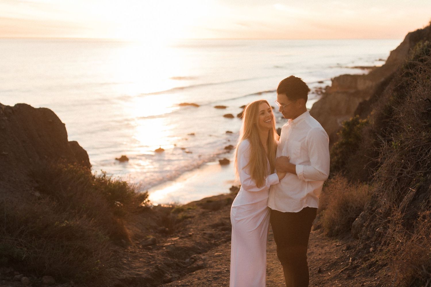 A romantic coastal sunset photoshoot with a couple in white attire against dramatic cliffs.