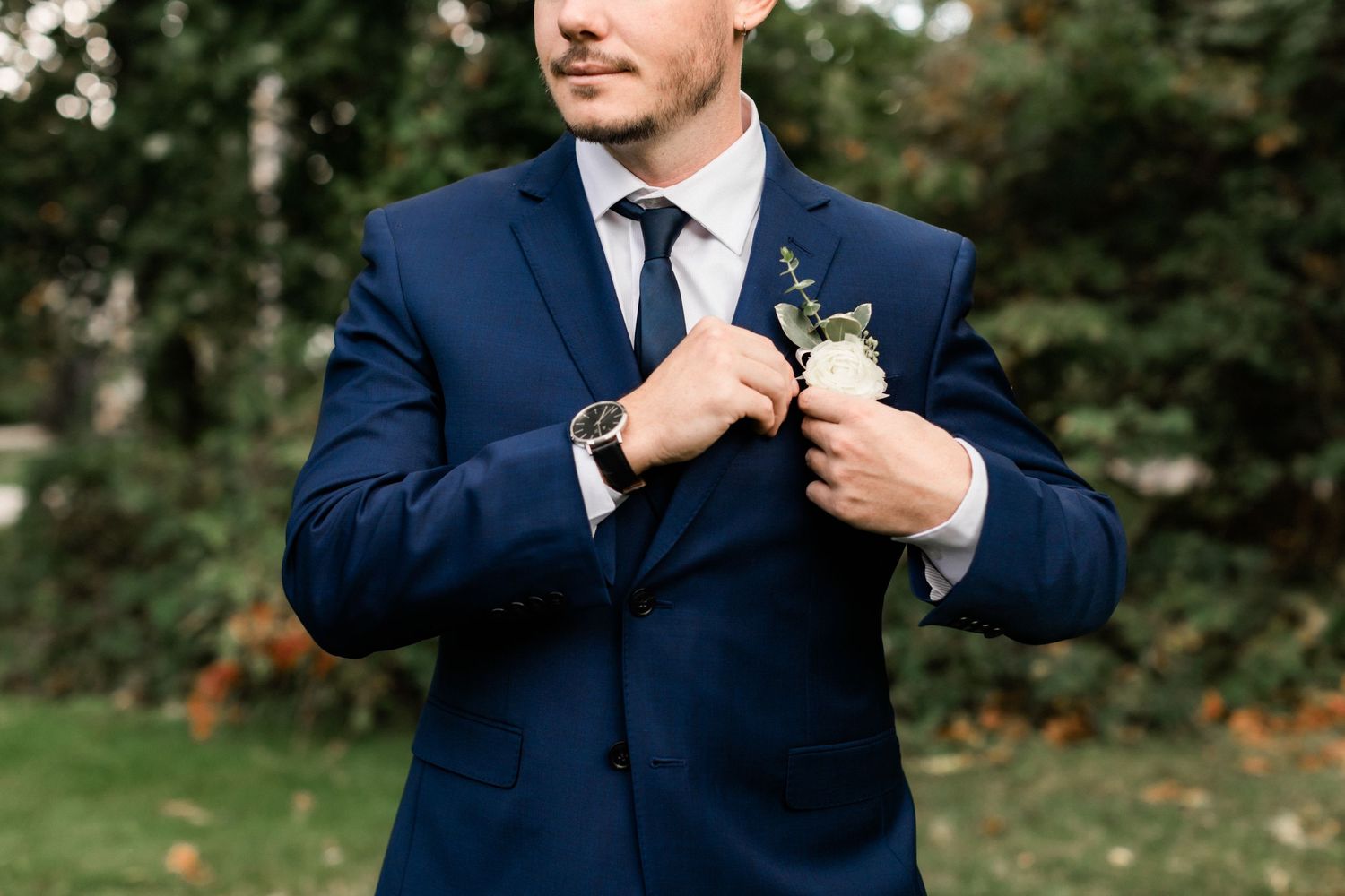 Groom in navy blue suit adjusts his boutonniere while standing outdoors.