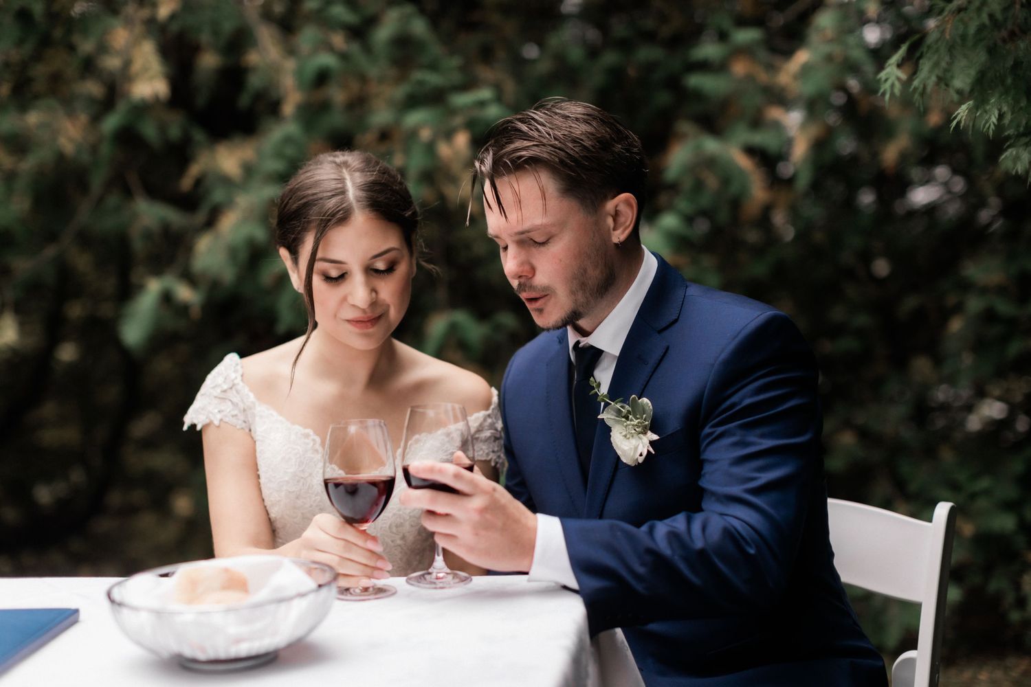 Outdoor wedding reception scene with couple seated at white-clothed table being served.