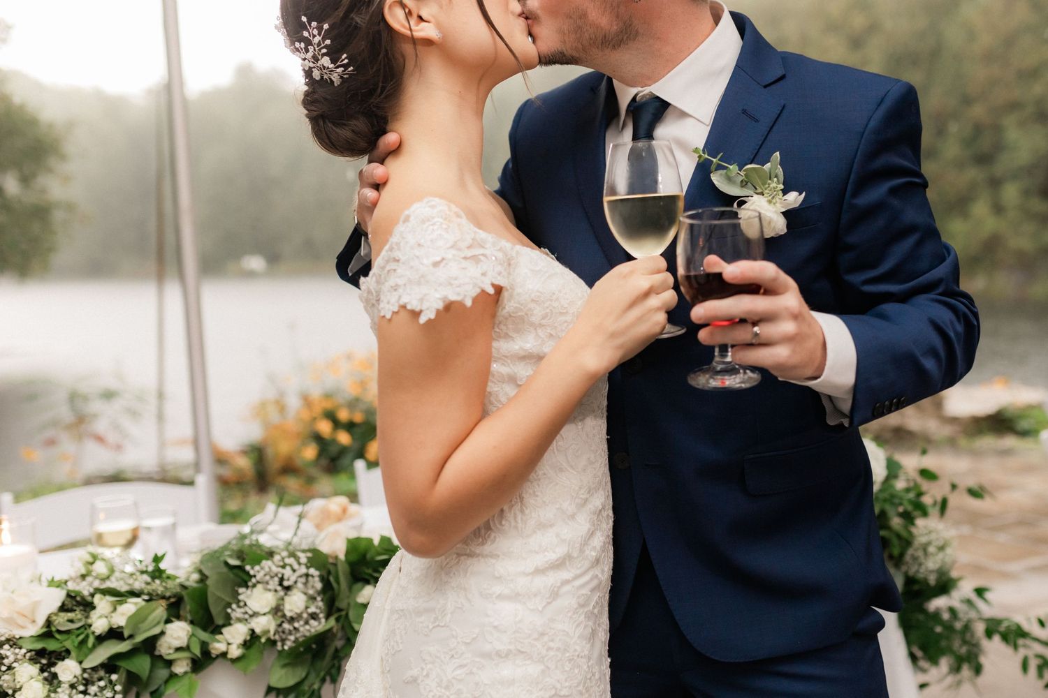 A wedding couple in classic attire share a tender moment with wine glasses.