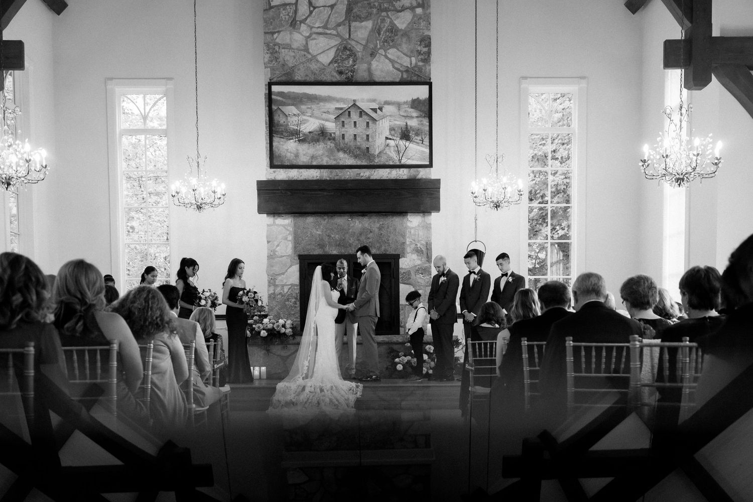 Black and white photo of a wedding ceremony in a chapel with guests seated in rows and chandelier lighting.