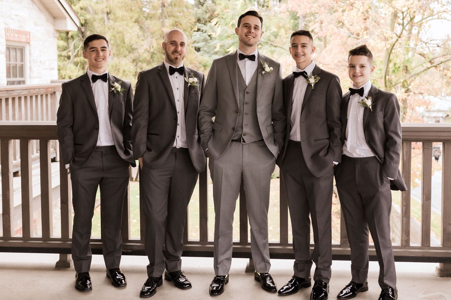 Five groomsmen in formal attire stand together on a covered porch.