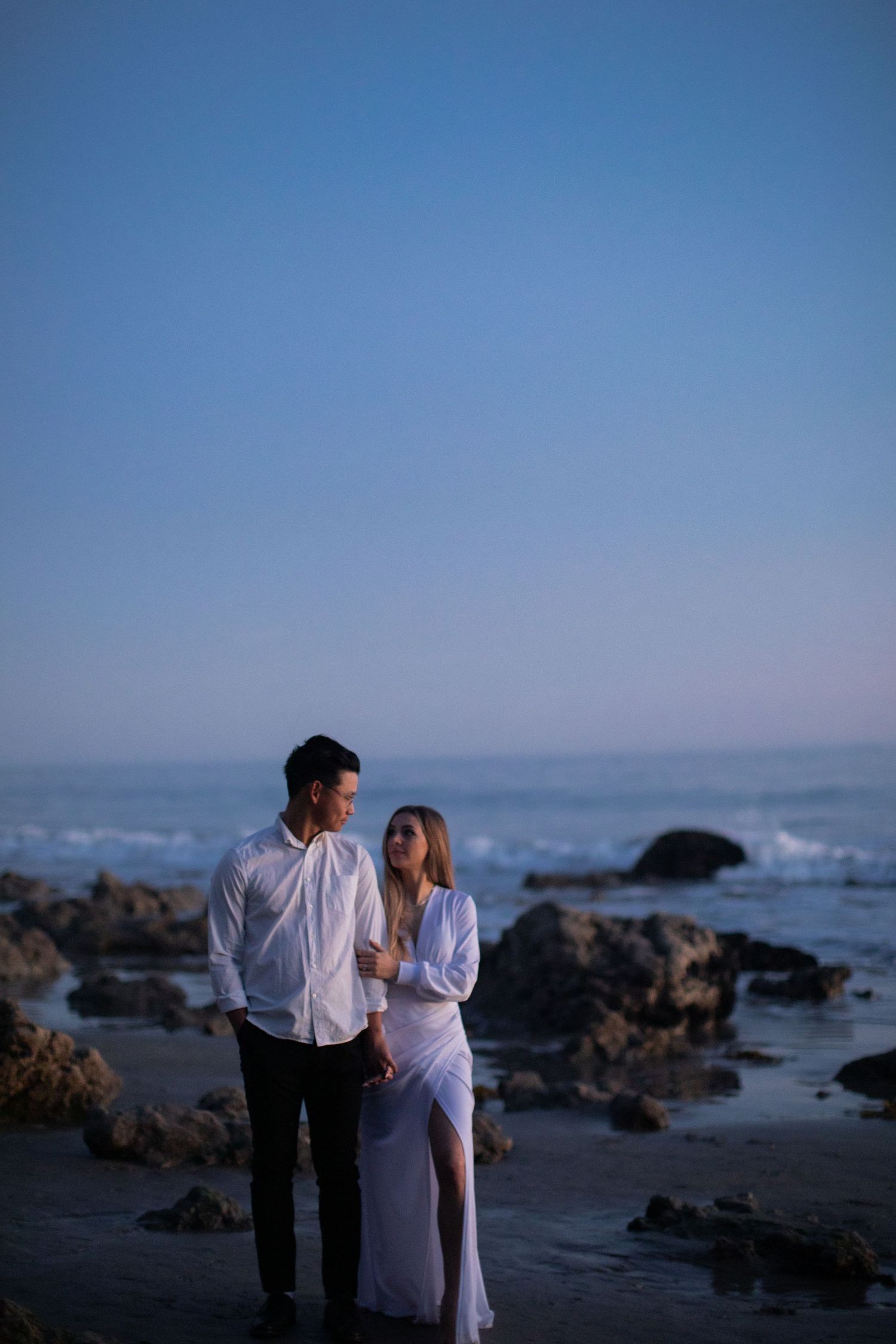 Romantic couple in white outfits embracing on rocky beach at dusk.