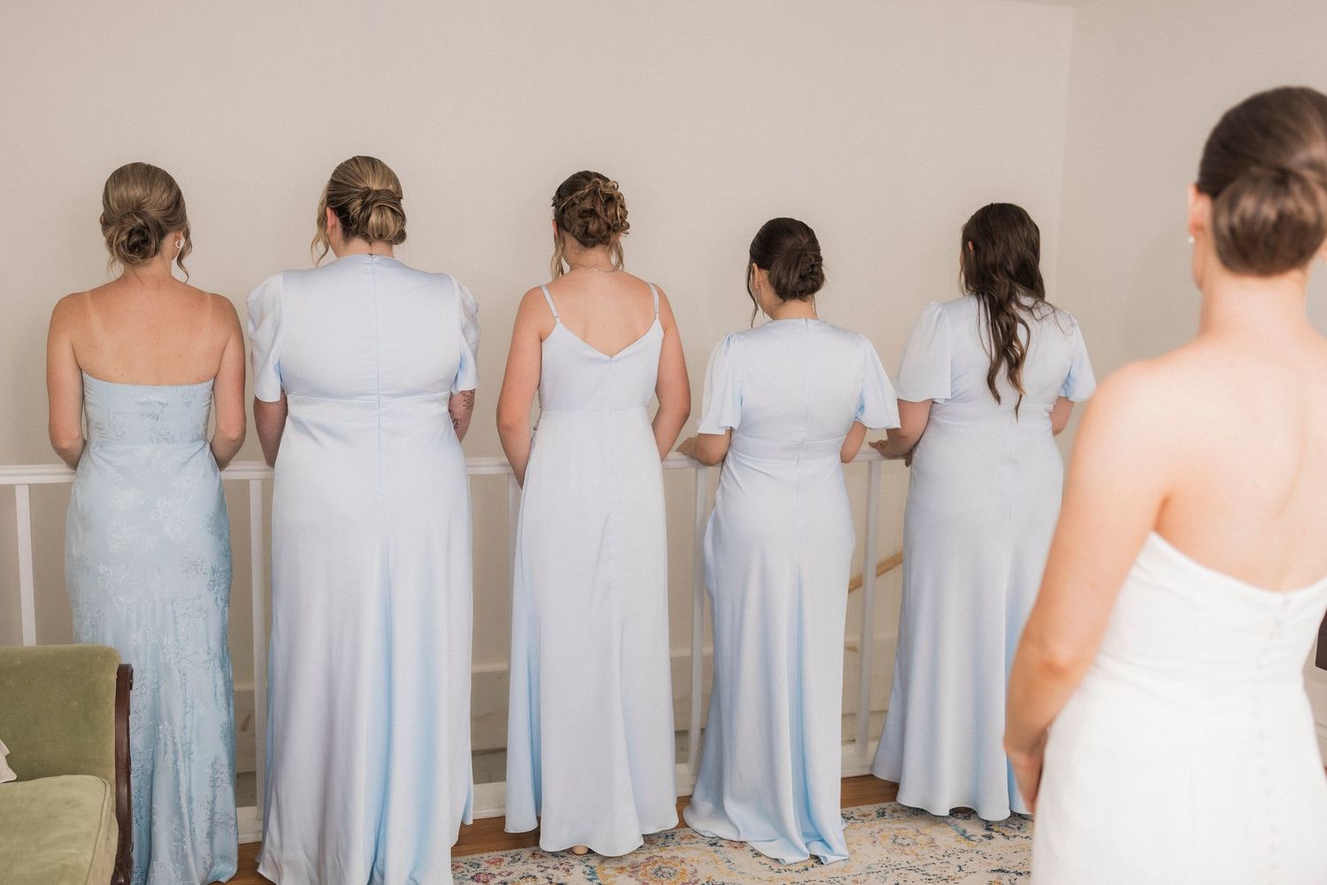 A lineup of bridesmaids in pale blue and white dresses standing together showing the backs of their gowns.