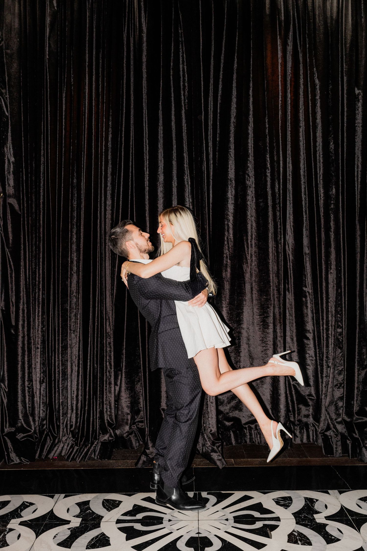 A couple in formal attire embrace against a dark curtain backdrop, with the woman in white lifting one leg playfully.