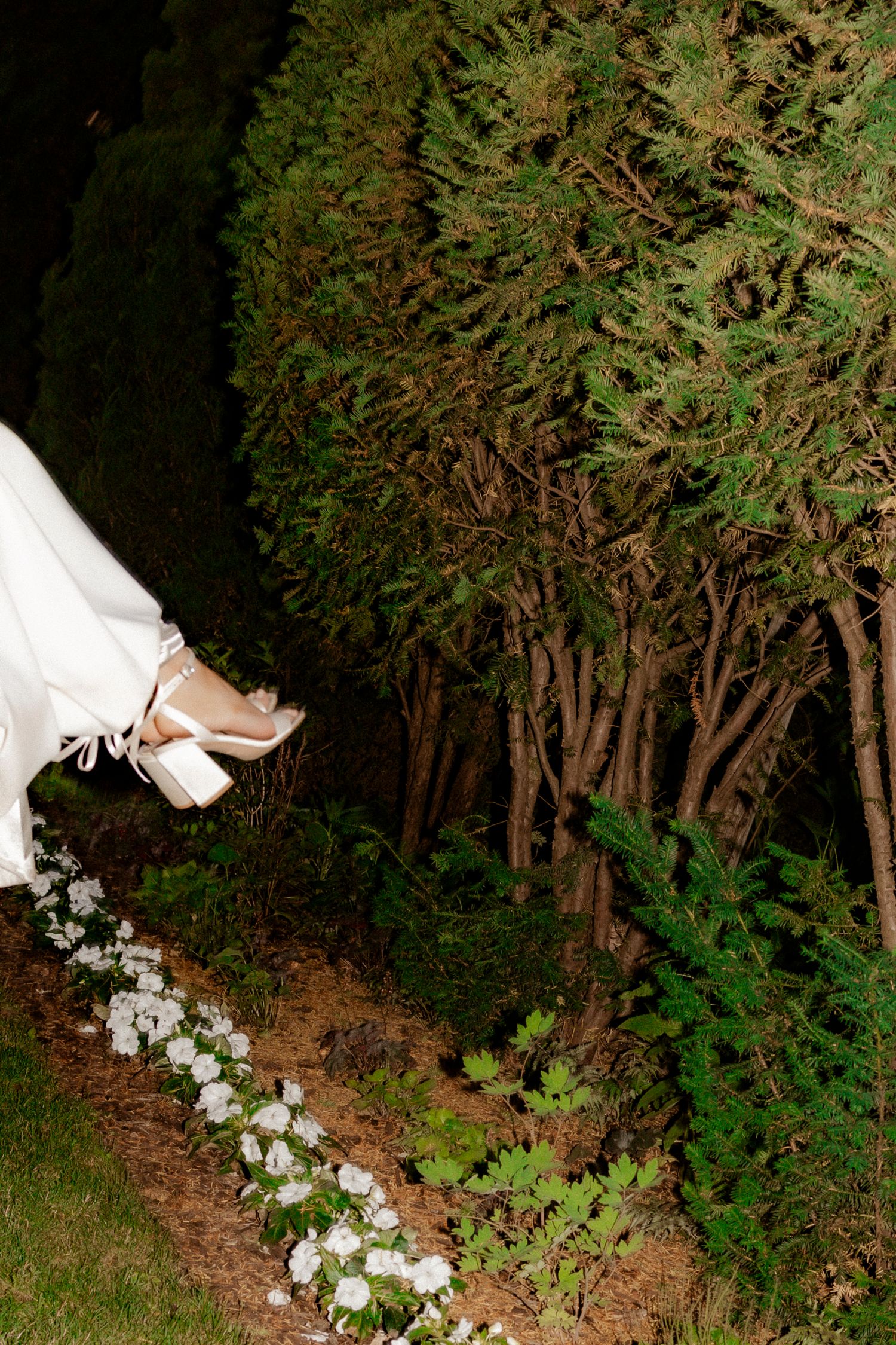 A white wedding dress and shoe next to a decorative floral arrangement on the ground against a backdrop of trees at night.