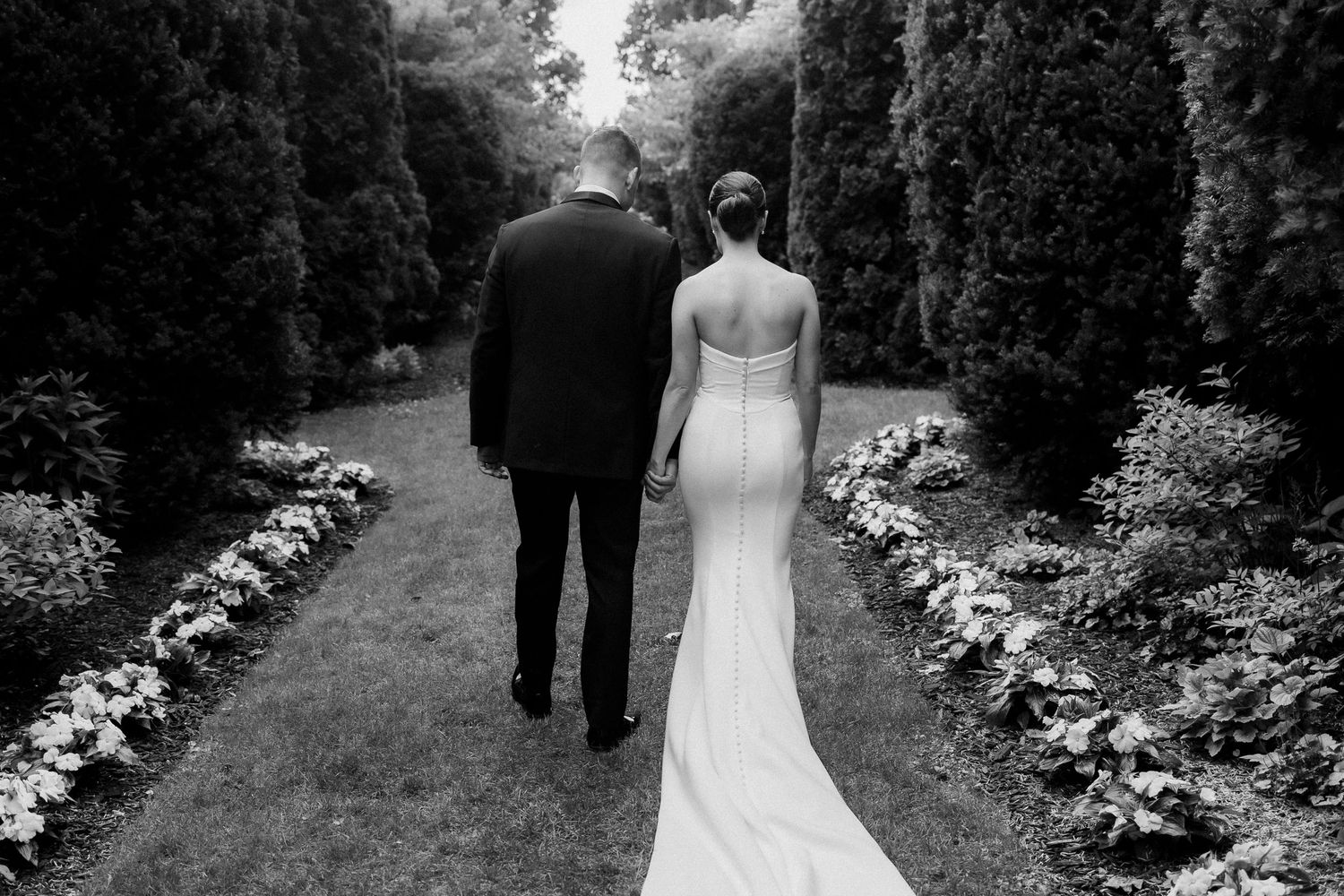 Black and white photo of couple walking down a garden path lined with flowers on their wedding day.