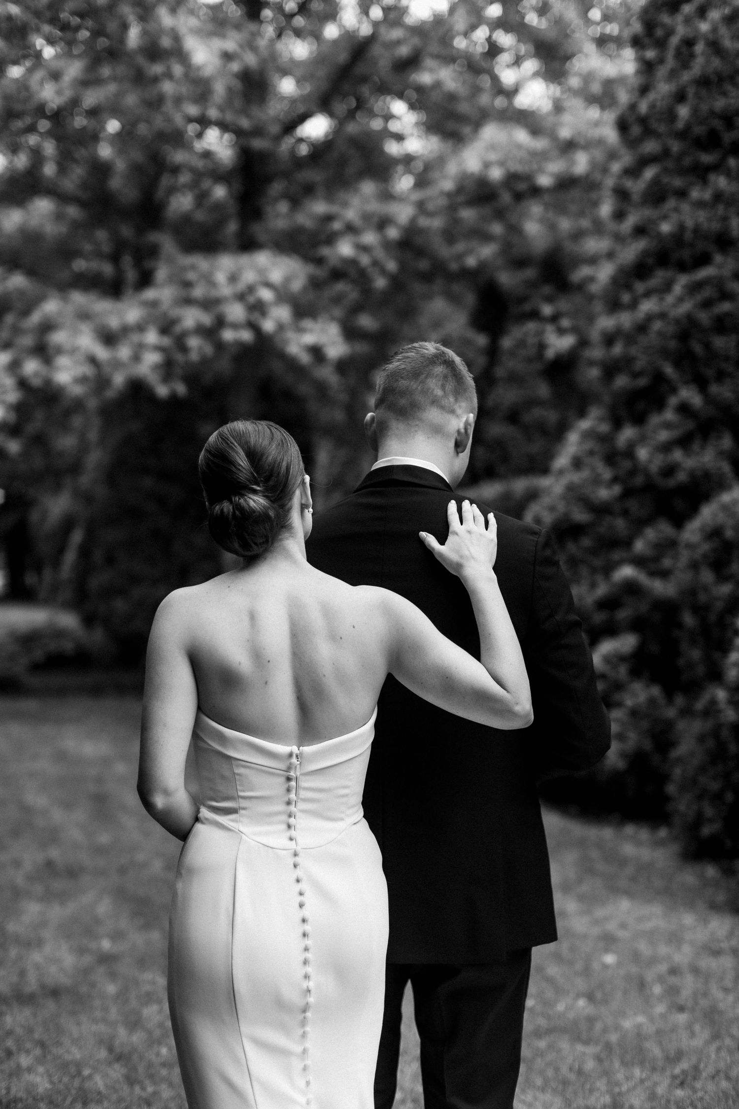 Black and white photo showing the back view of a bride and groom standing together in a garden setting.