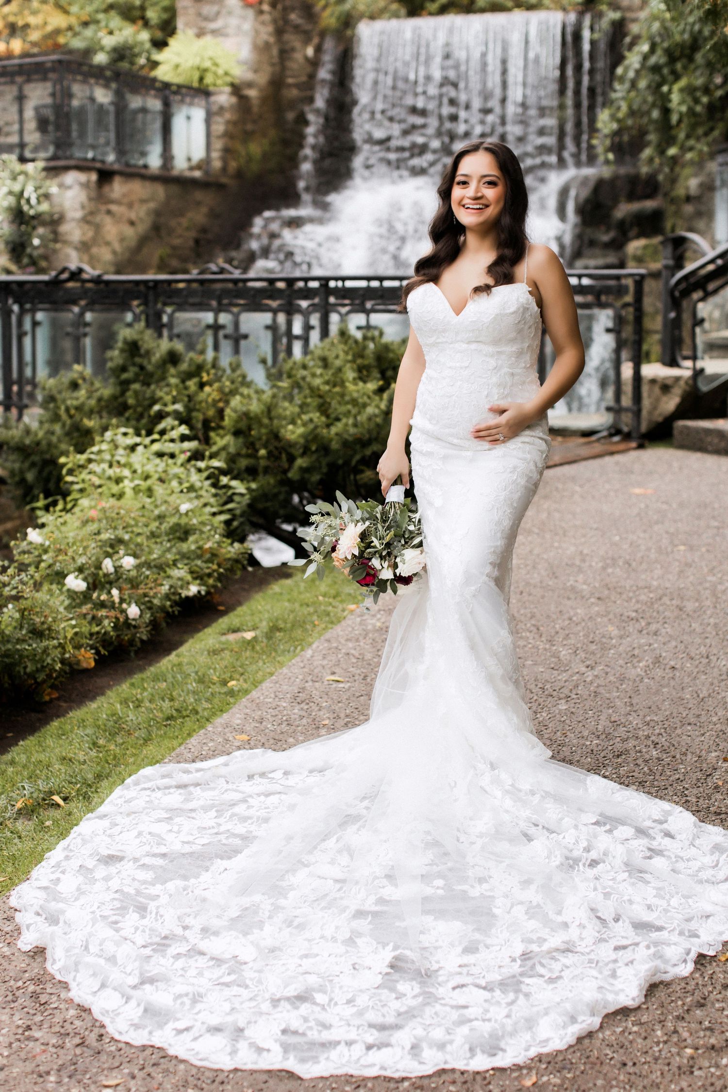 Elegant bride in lace mermaid wedding dress with long train poses by scenic waterfall.
