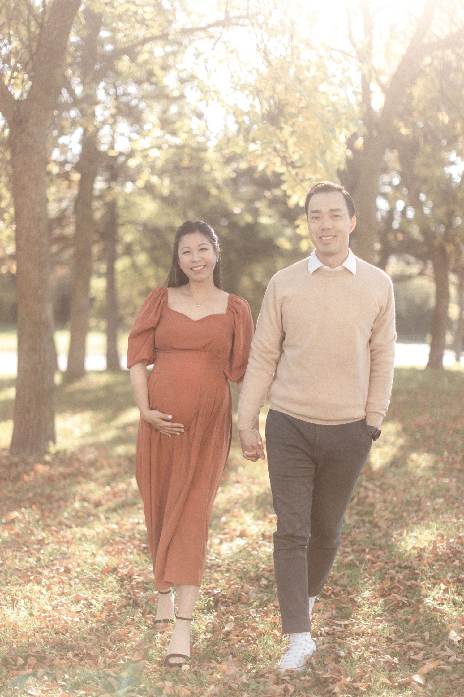 A couple in autumn attire walks hand in hand through a park with golden sunlight streaming through trees.