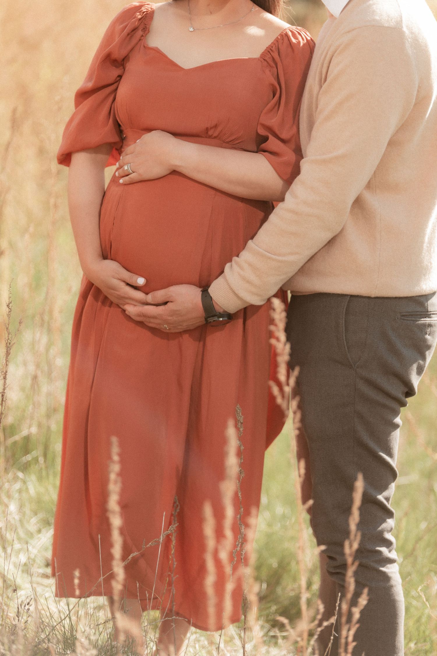 Close-up of pregnant woman in rust dress with partner's hands on her belly in wheat field.