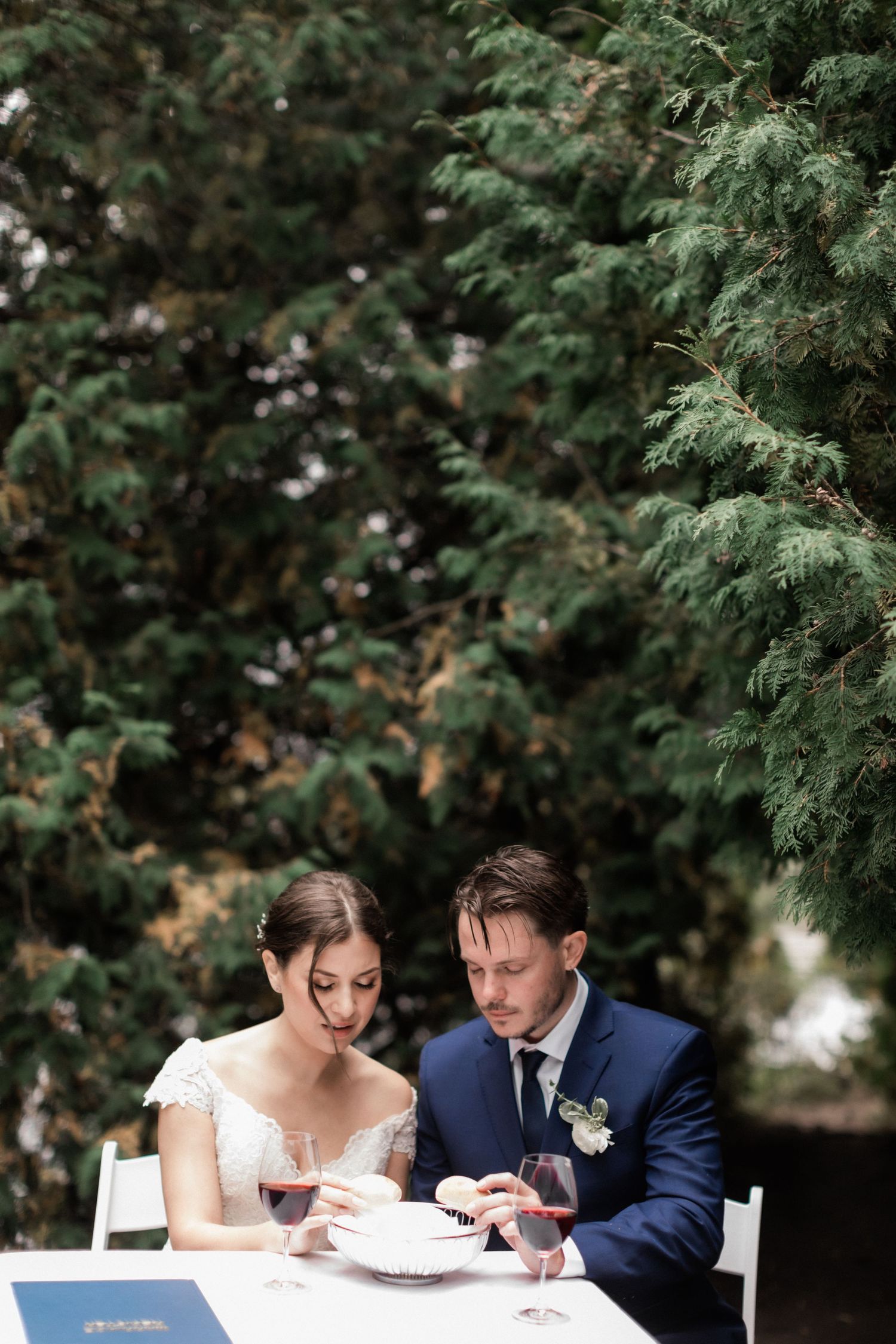 Couple sits at outdoor table during intimate forest wedding ceremony with wine glasses.
