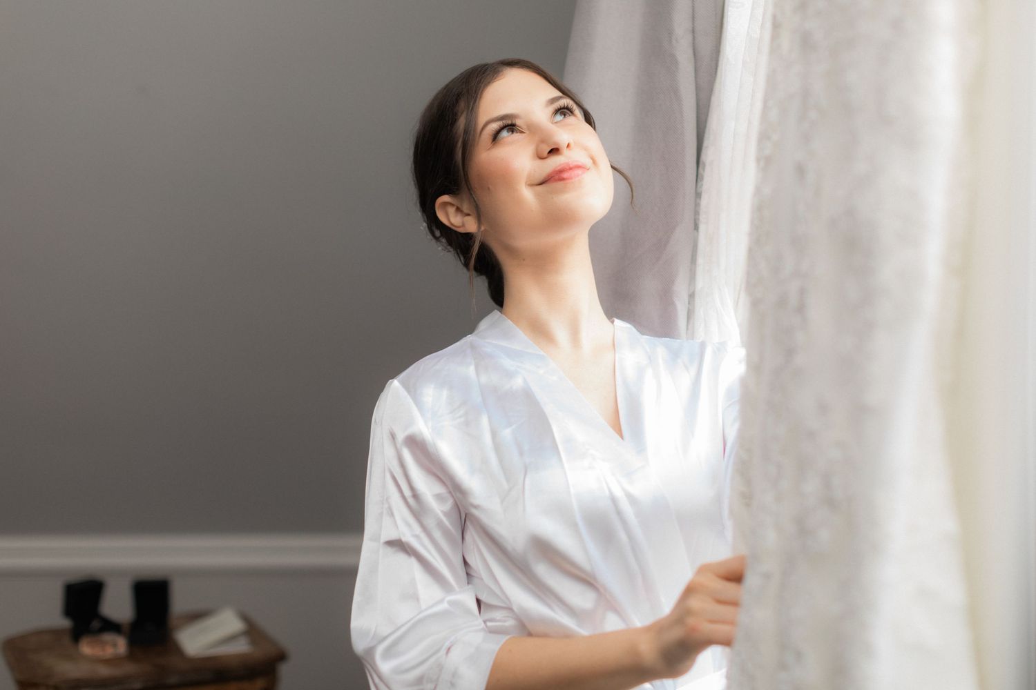 Person in white satin robe gazes up while standing near window.
