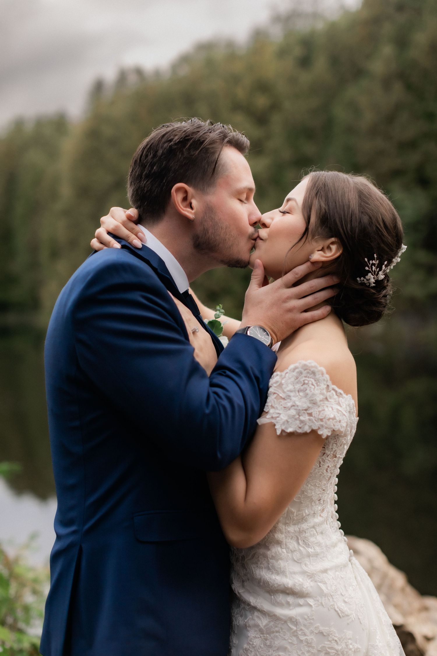 Romantic lakeside wedding kiss with evergreen forest in background.