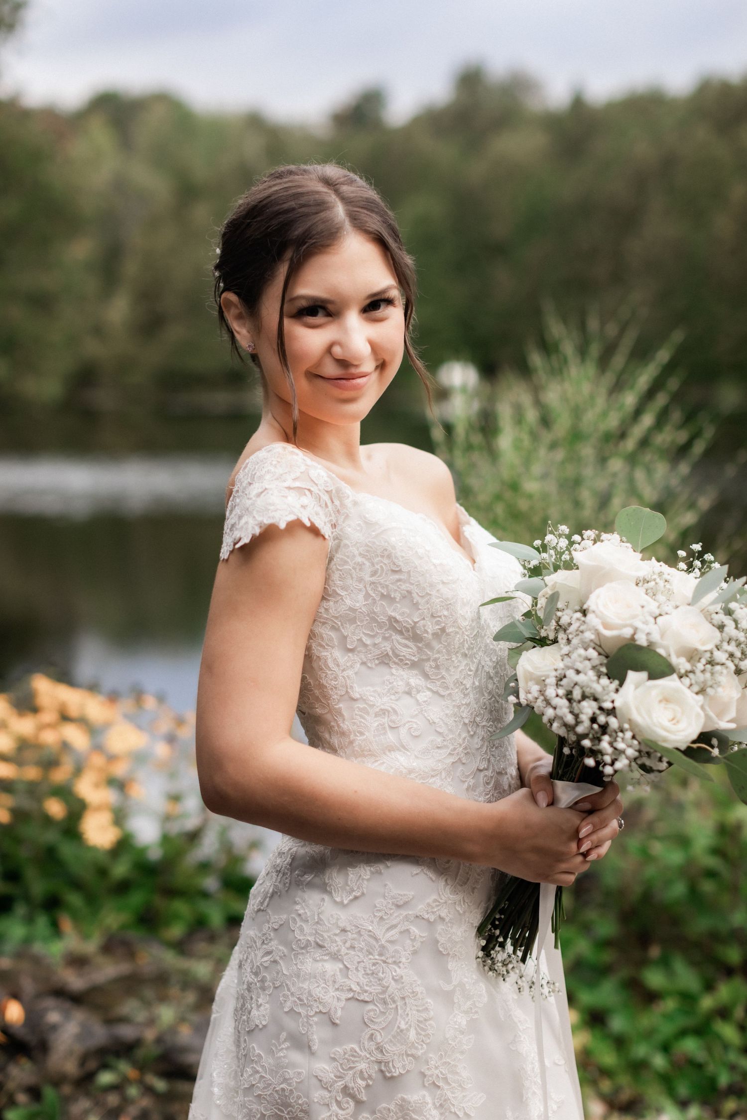 Bride poses with white bouquet in lace dress against natural landscape with lake.