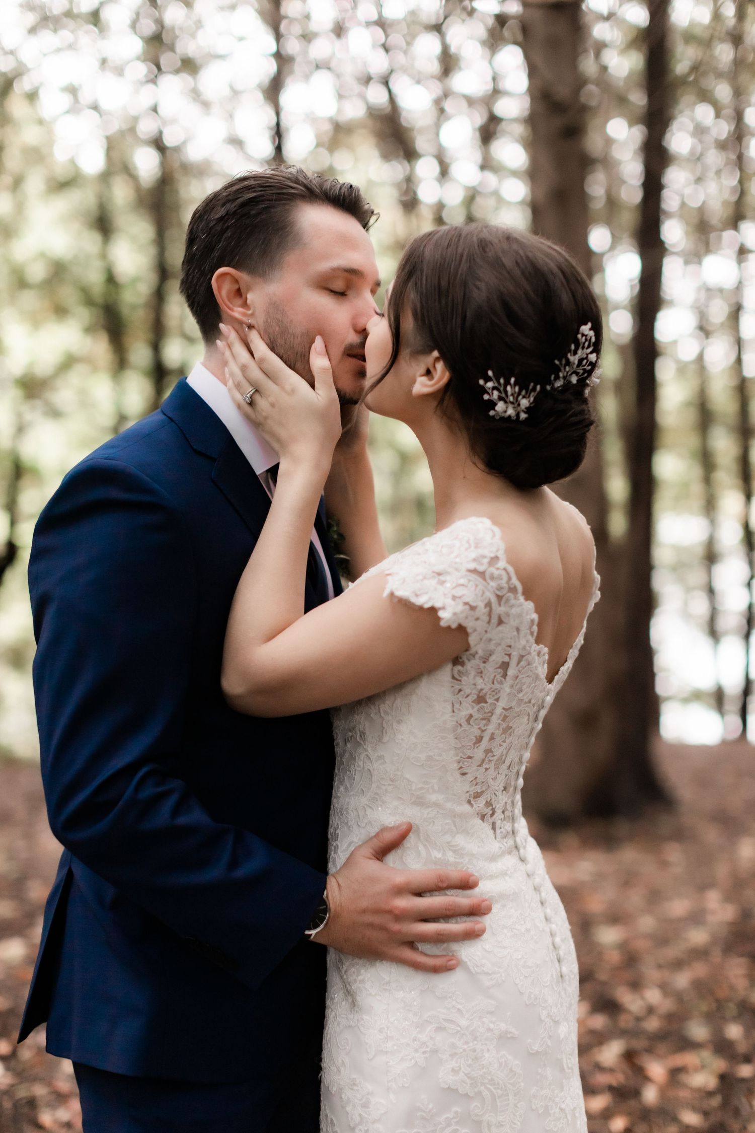 Intimate wedding portrait in forest with bride in lace gown and groom in navy suit sharing tender moment.