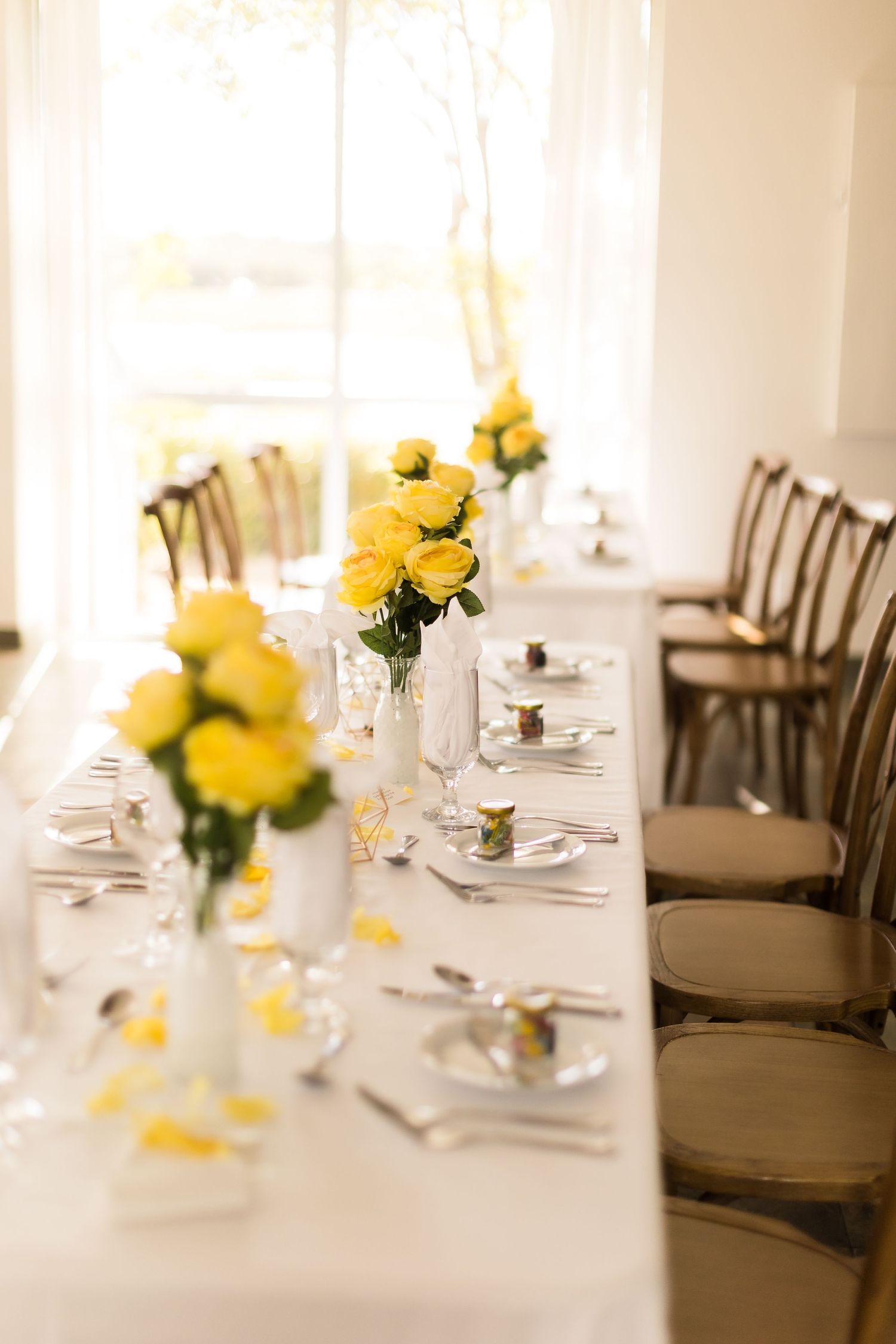 Elegant wedding table setting with yellow rose centerpieces in white vases and wooden chairs.