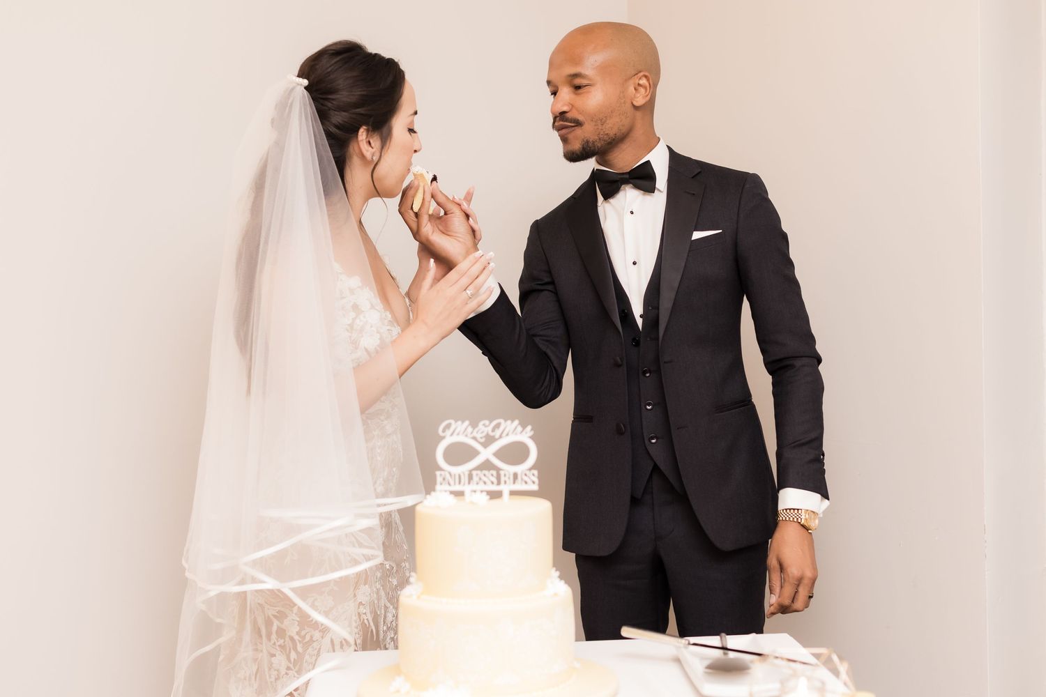 A bride and groom share a playful moment while cutting their elegant white wedding cake with an infinity symbol topper.