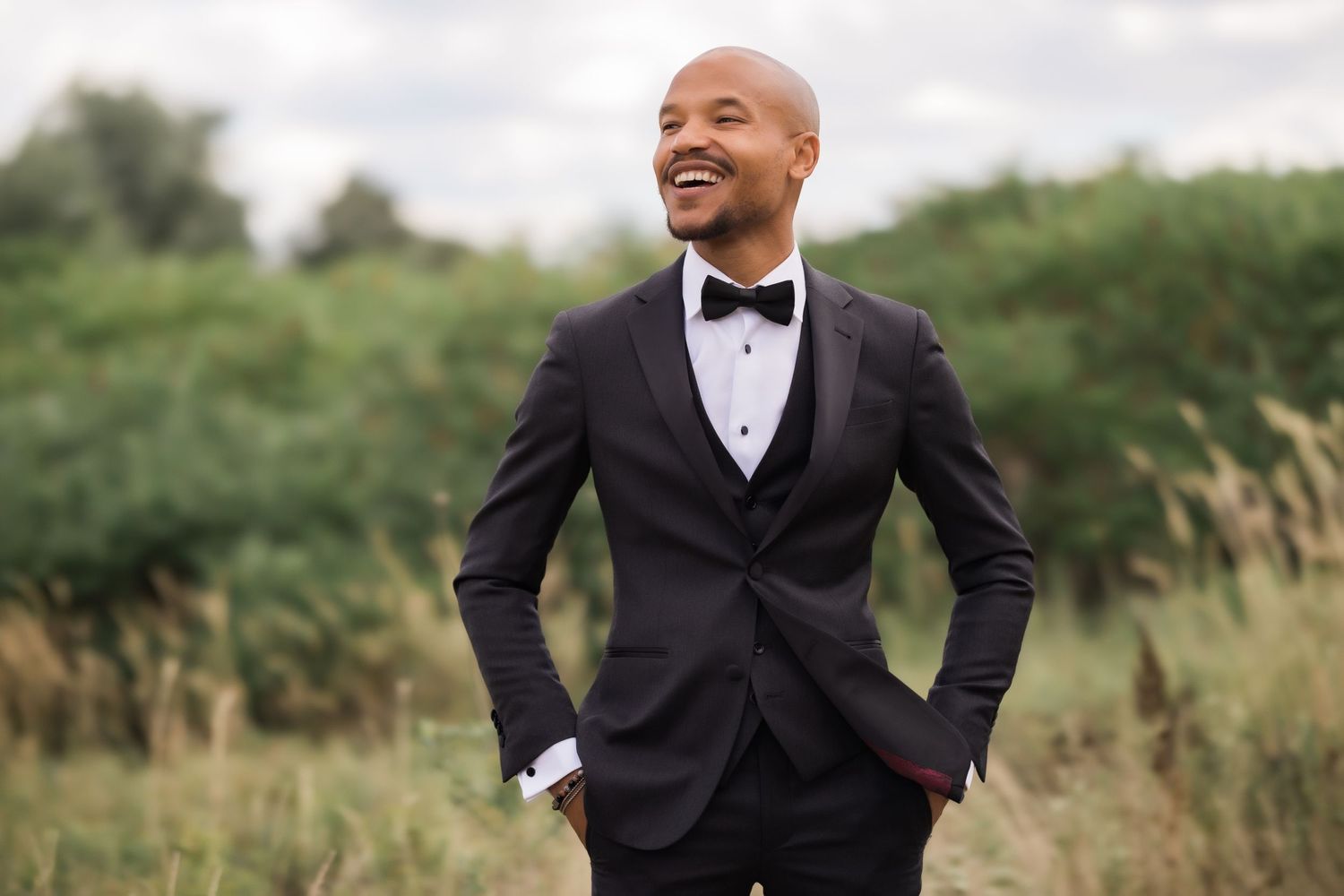 Groom in classic black tuxedo smiles while posing in outdoor field setting.