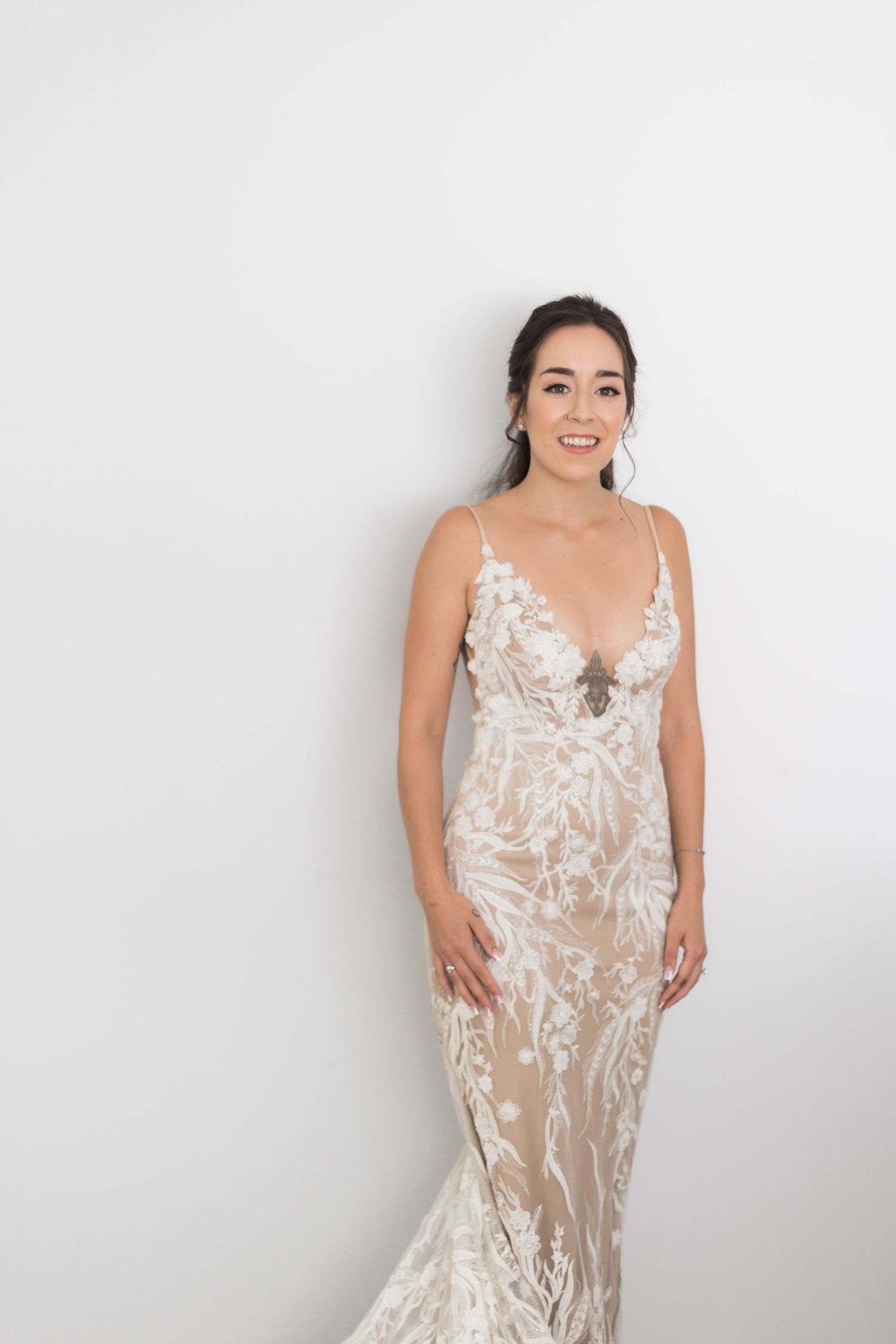 Bride wearing elegant white lace wedding gown poses against white wall.