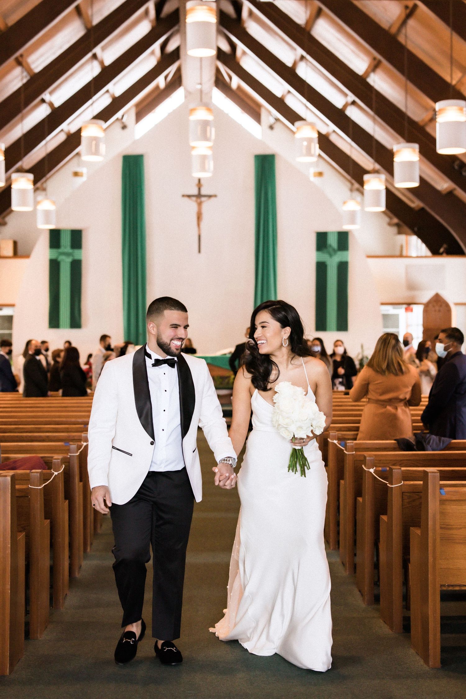 Newlyweds smile while walking hand in hand down church aisle with cross in background.