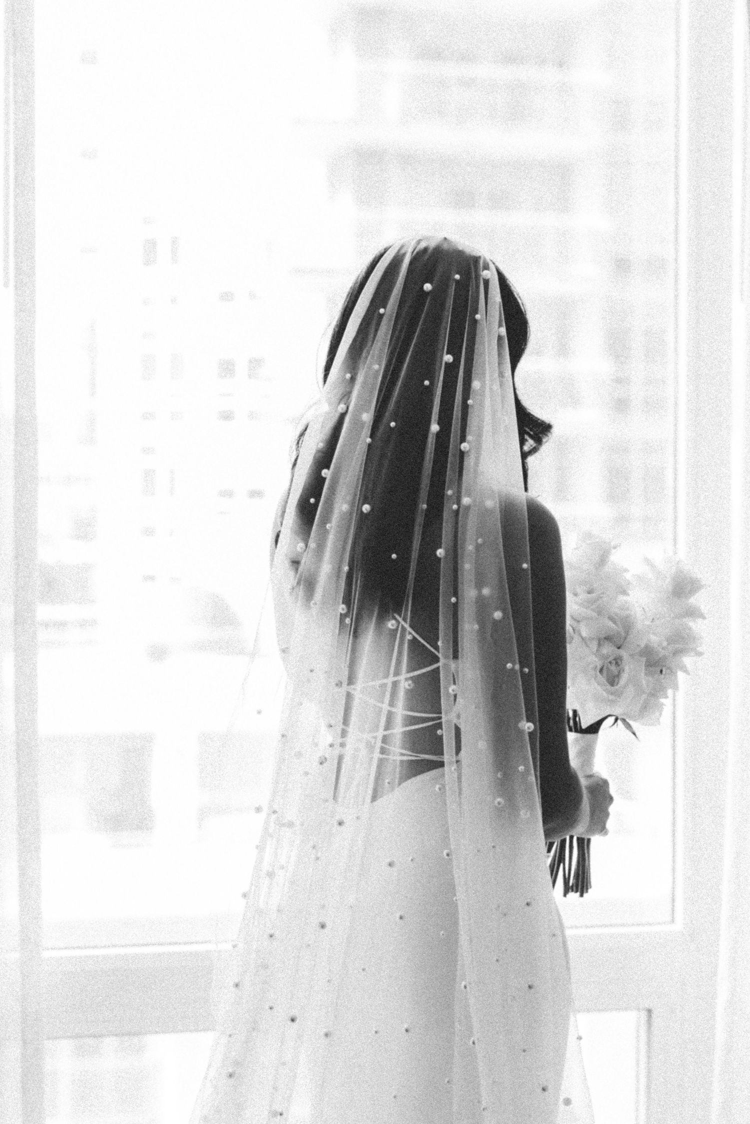 Silhouette of bride with sparkling veil standing by window holding bouquet.