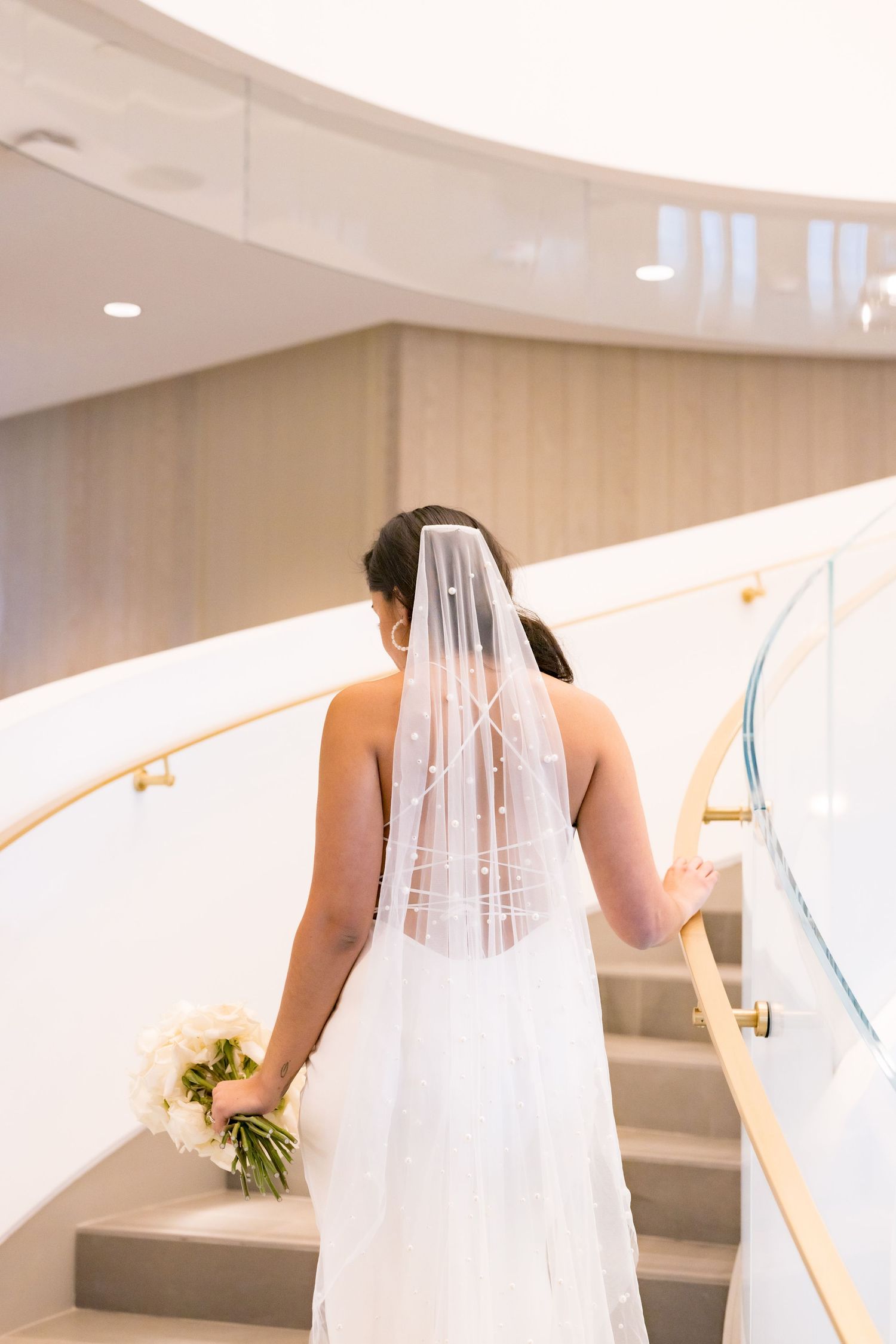 A bride in a white wedding dress and veil climbs a modern staircase while holding a bouquet.