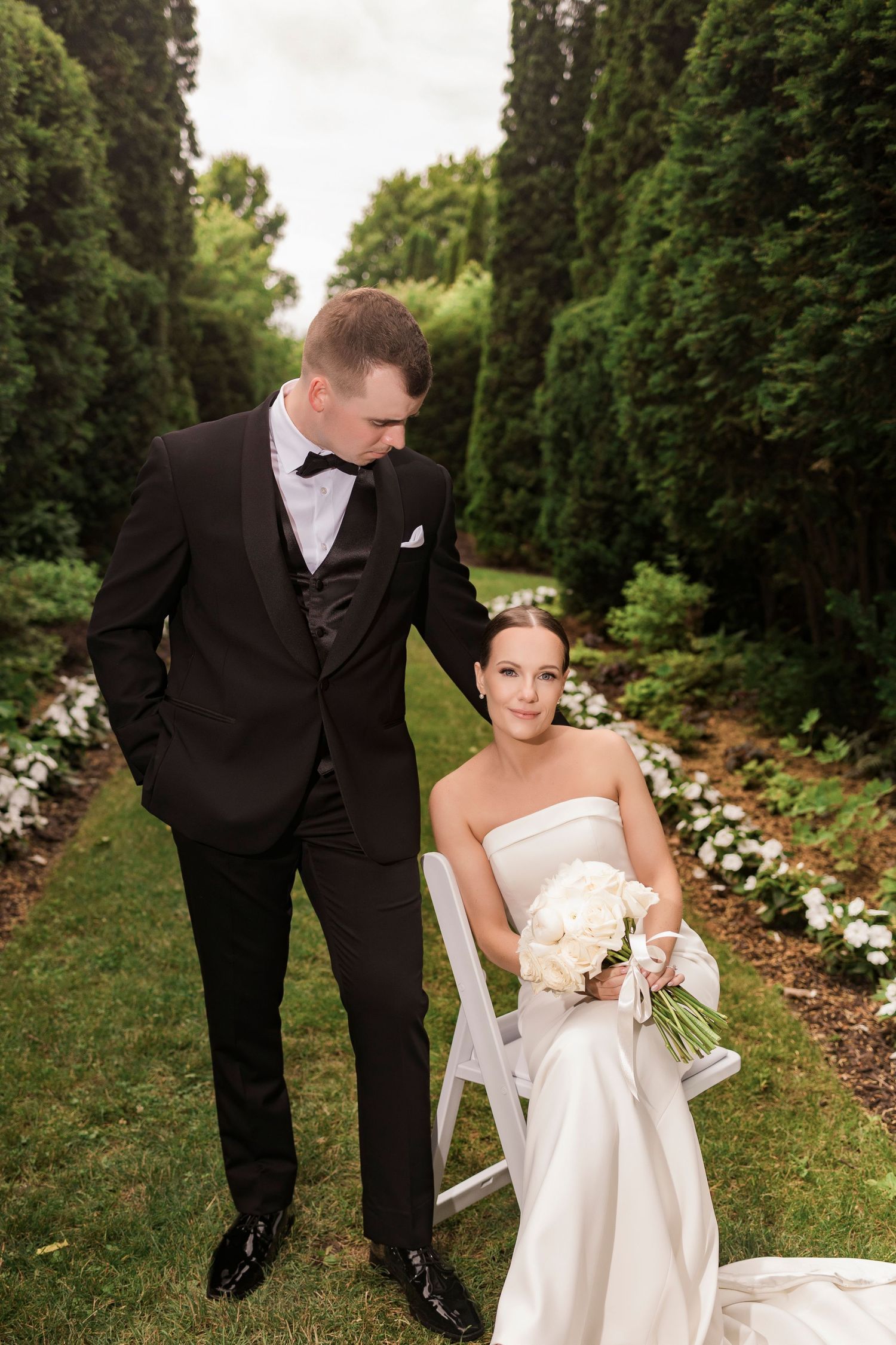 Elegant couple in wedding attire pose together on flower-lined garden path.