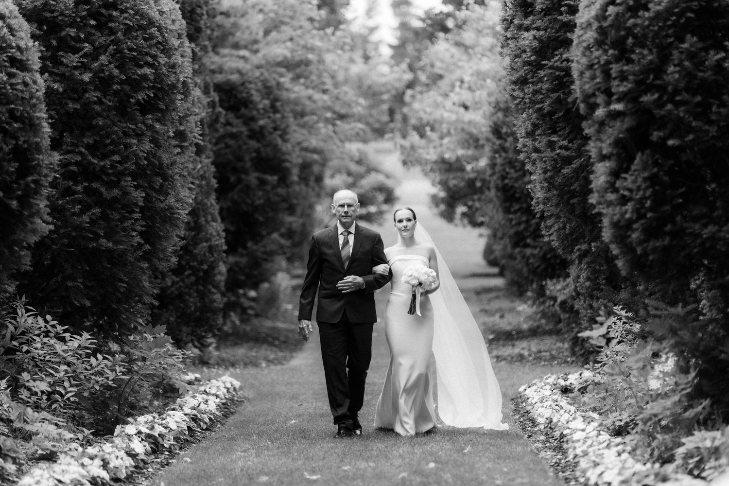 A black and white photograph of a couple walking down a garden path lined with tall hedges on their wedding day.
