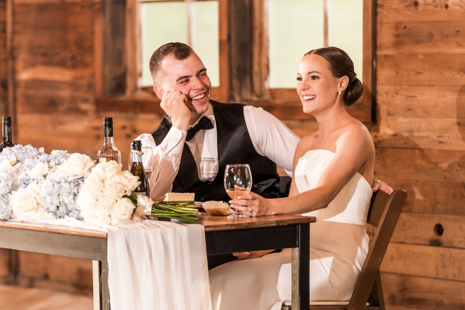 A newlywed couple shares a joyful moment while dining together at a rustic wooden table with floral decorations.