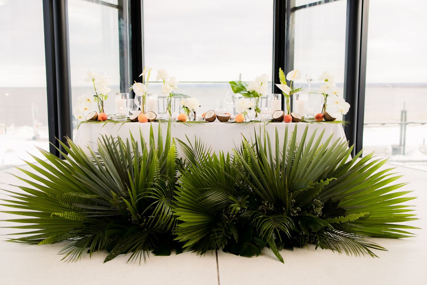 Large palm frond arrangements decorating white dining table in modern event space.
