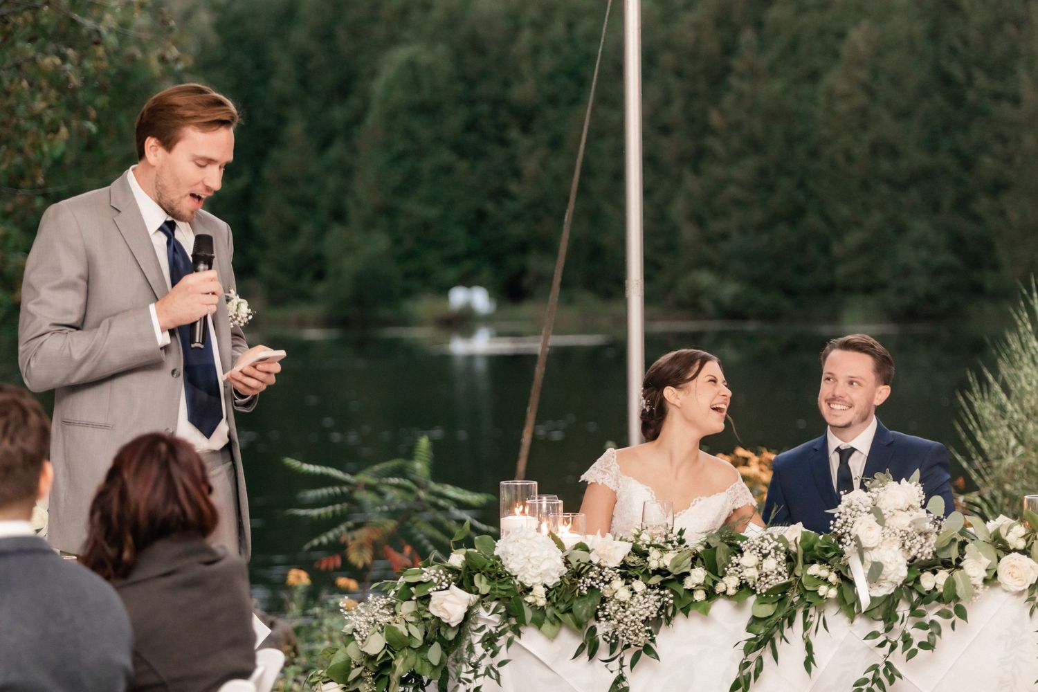 Wedding toast being given during outdoor reception by lakeside with floral table decorations.