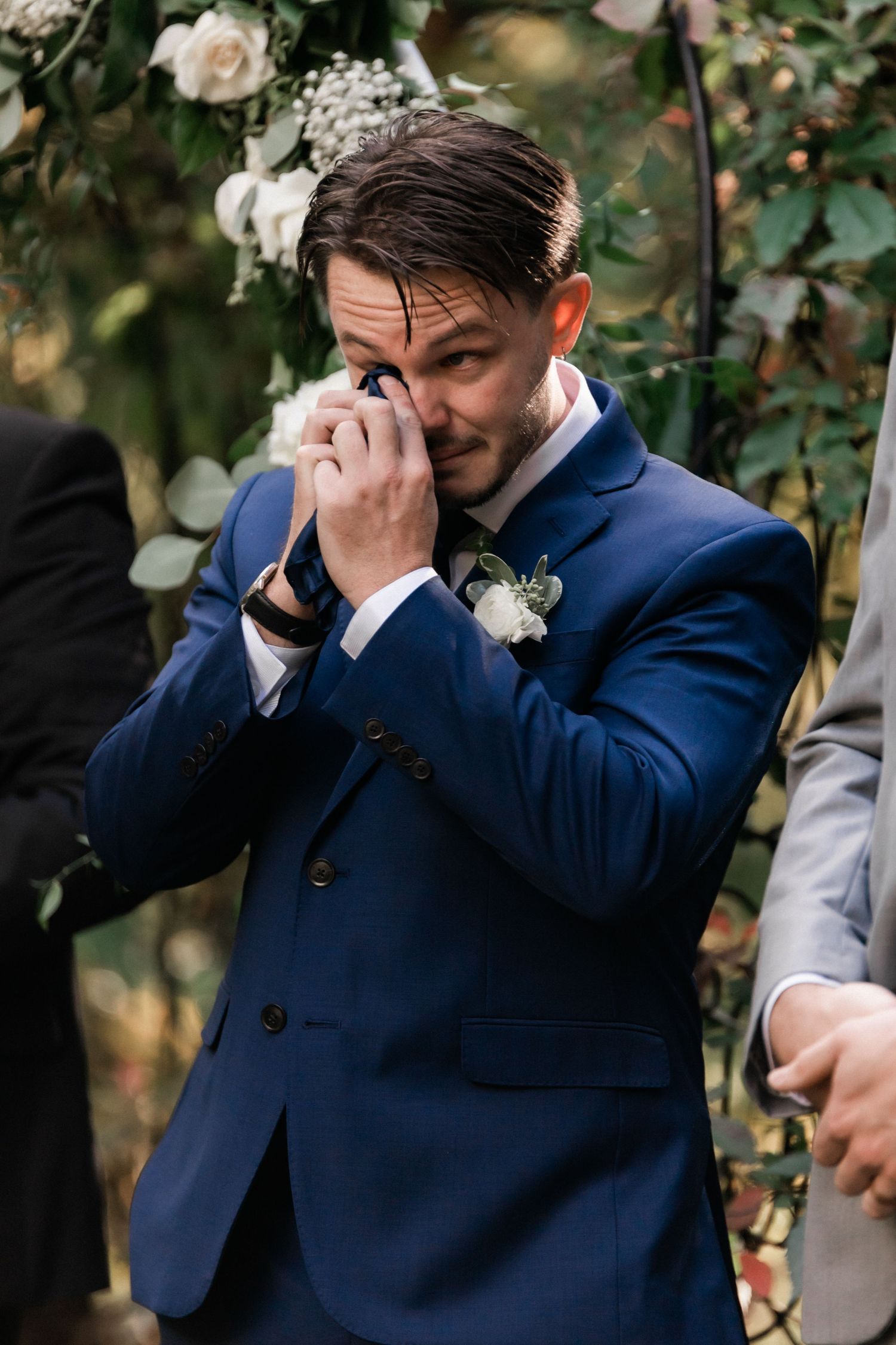 An emotional groom wipes tears from his eyes during an outdoor wedding ceremony.