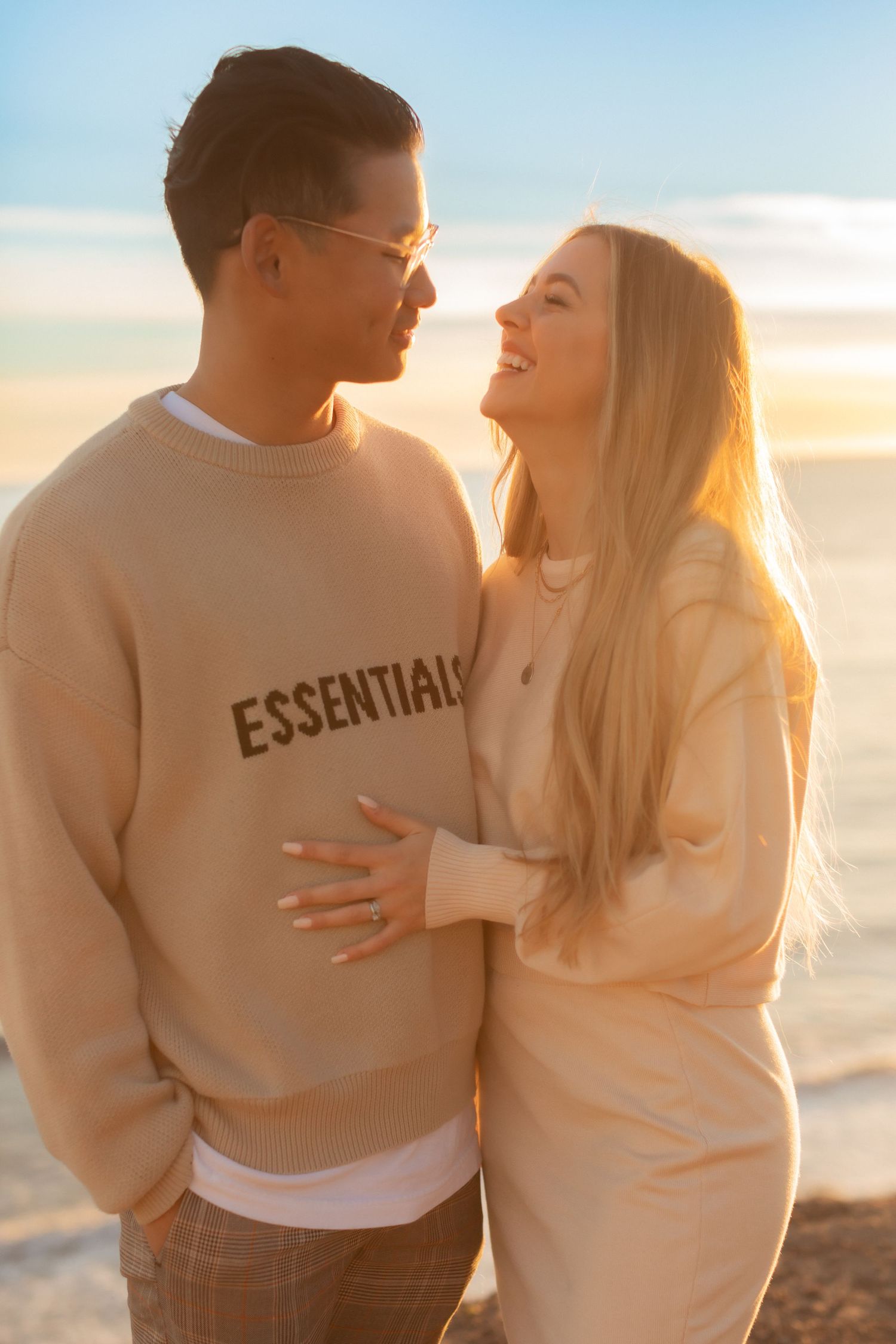 Couple in cream sweaters sharing loving moment against sunset ocean backdrop.