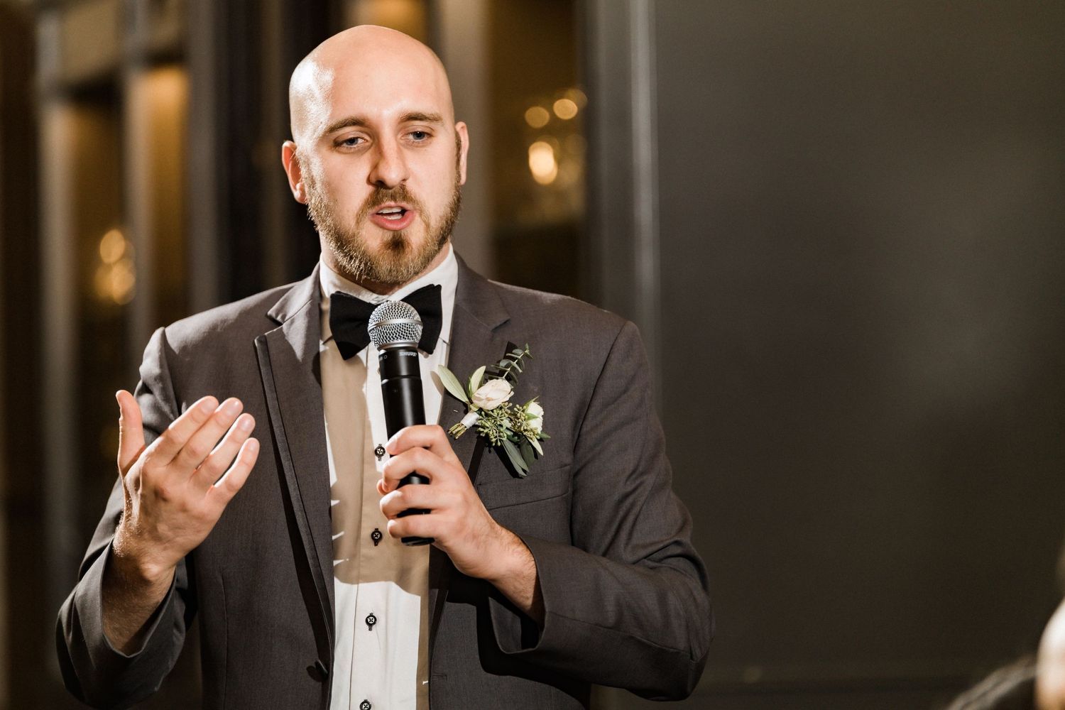 A person in grey suit gives a speech at formal event with microphone and boutonniere.