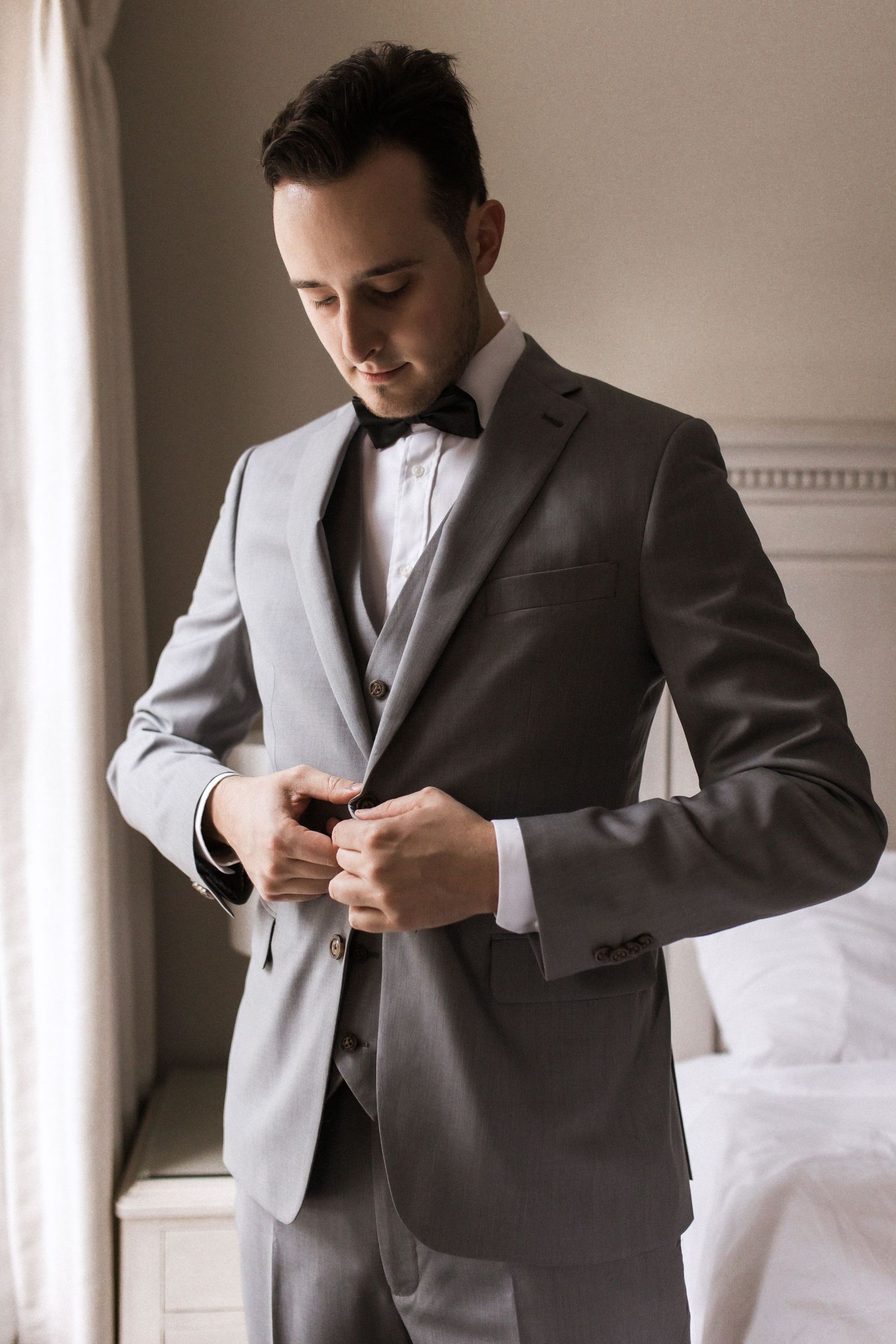 Groom in grey suit buttons jacket while getting ready by window.