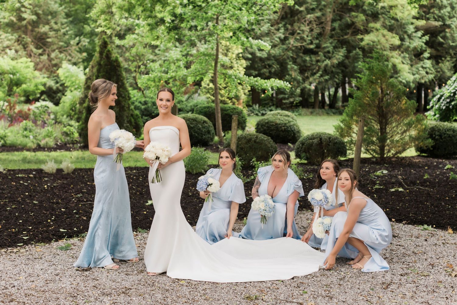 Bridesmaids in light blue dresses gather around bride on garden grounds during wedding photoshoot.