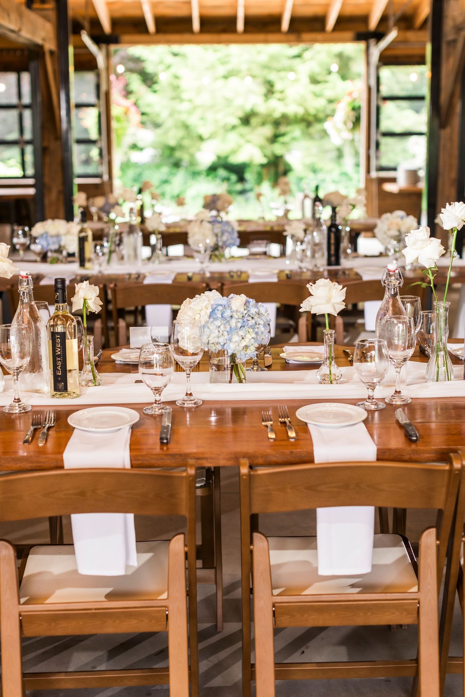 Rustic wooden tables and chairs set for a wedding reception with elegant glassware and table settings in a covered venue.