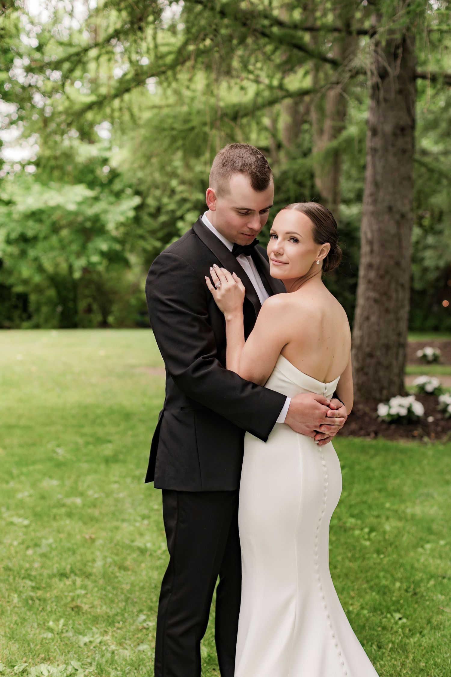 Elegant couple in wedding attire embrace on a manicured lawn with trees in background.