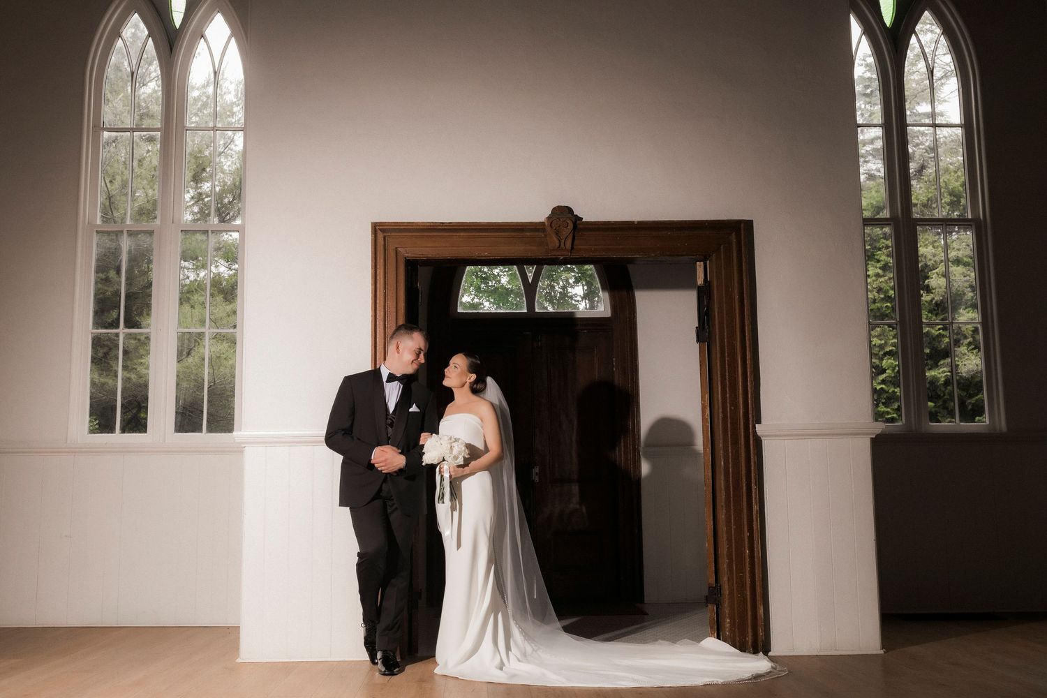 A couple stands in an elegant white chapel doorway framed by Gothic-style windows during their wedding ceremony.