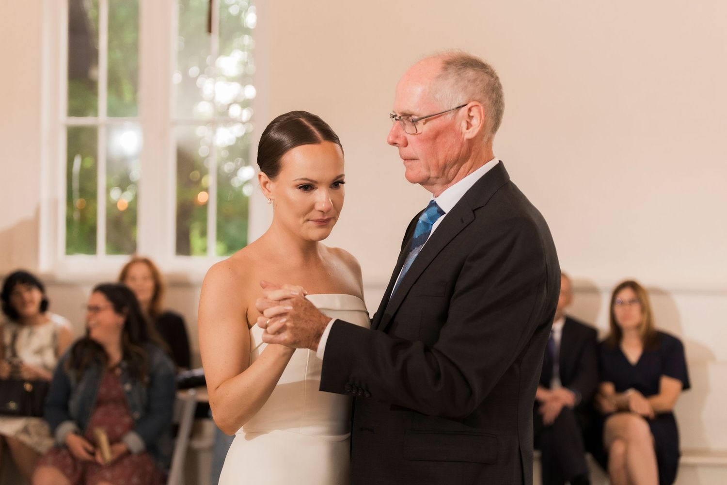 Emotional dance captured during wedding reception in bright indoor venue with large windows.