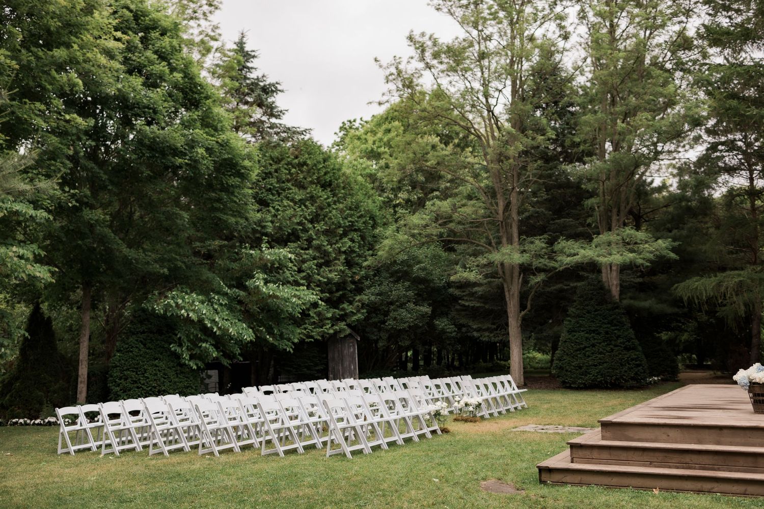White folding chairs arranged in rows for an outdoor wedding ceremony in a wooded garden setting with wooden steps.