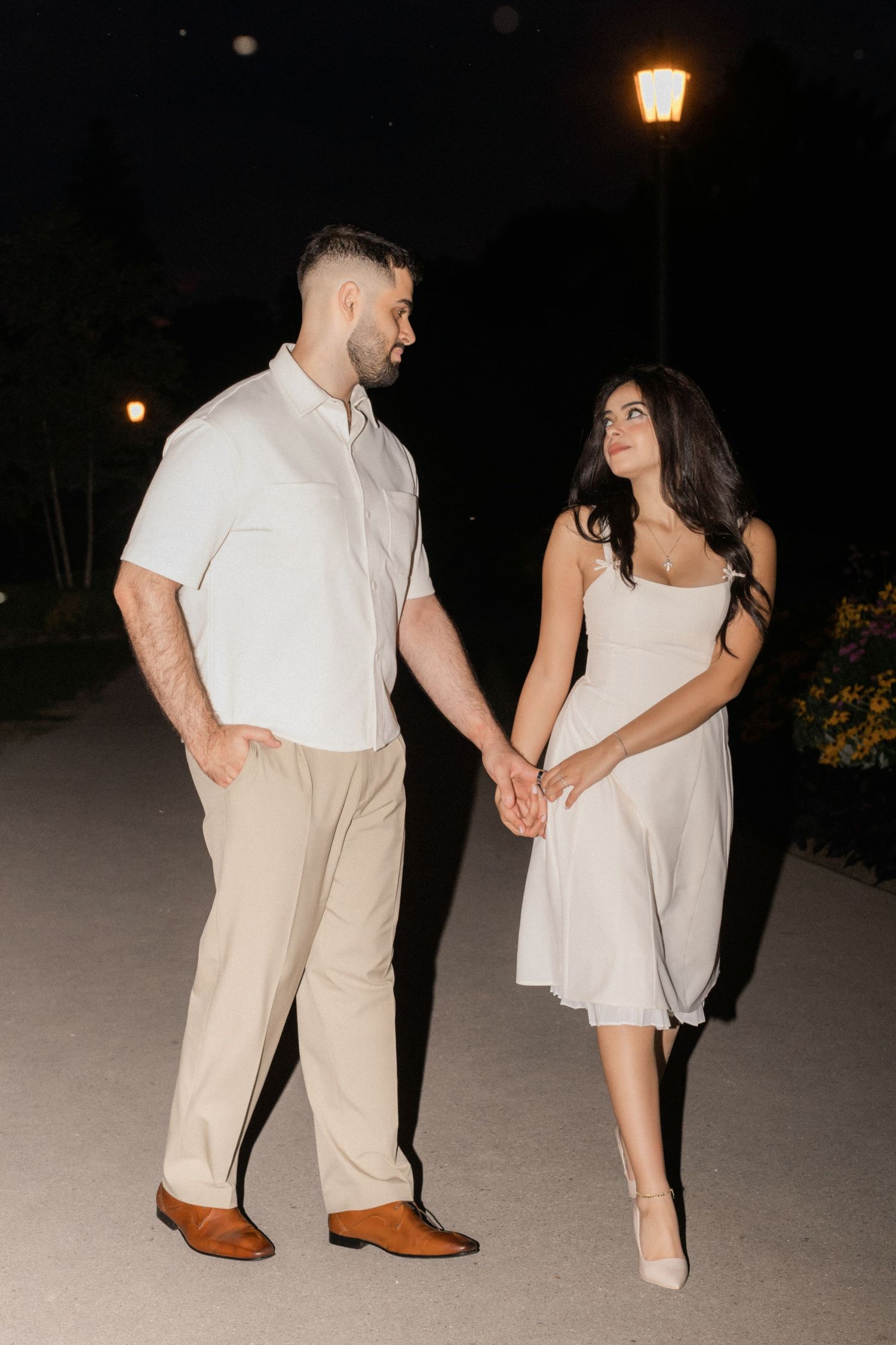 A couple in light colored outfits holds hands while taking an evening stroll under a glowing street lamp.