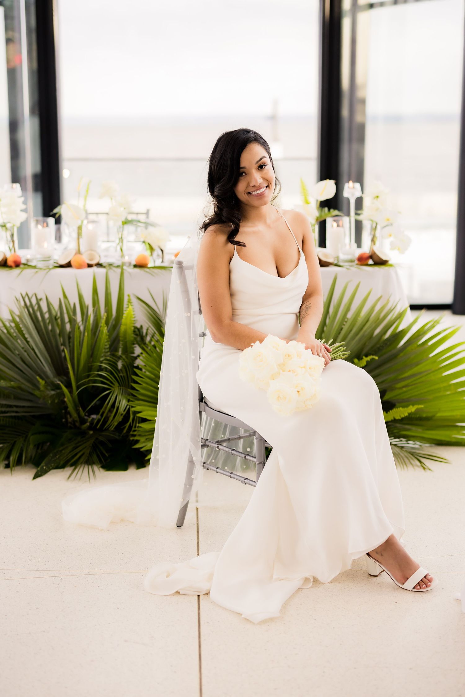 A bride in a white wedding dress sits elegantly on a silver chair with tropical palm leaves in the background.