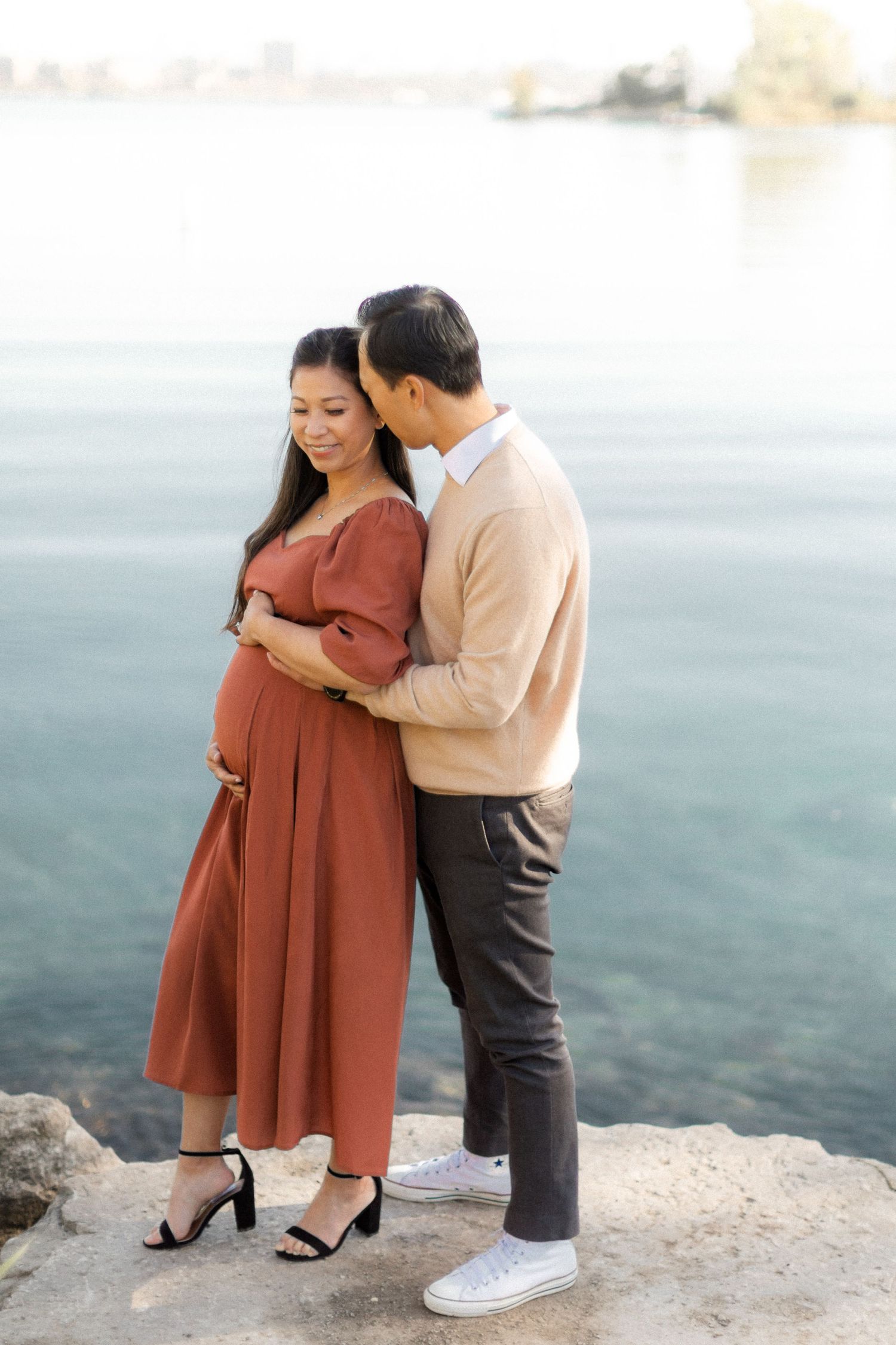 Expecting couple poses on rocky lakeside, dressed in beige sweater and rust dress.