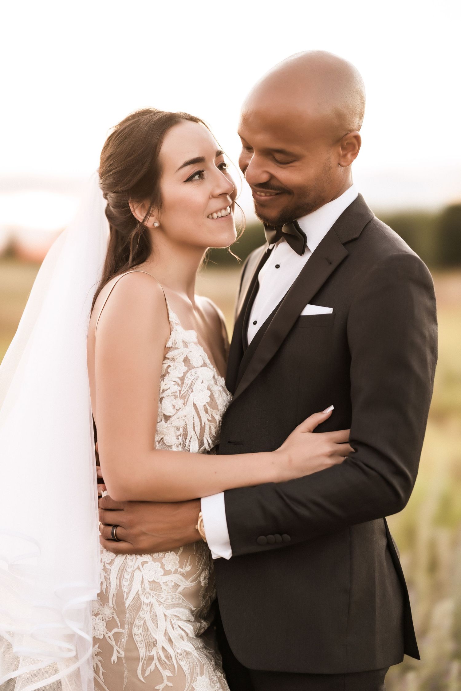 Bride in lace gown and groom in tuxedo share loving smiles during sunset portraits.