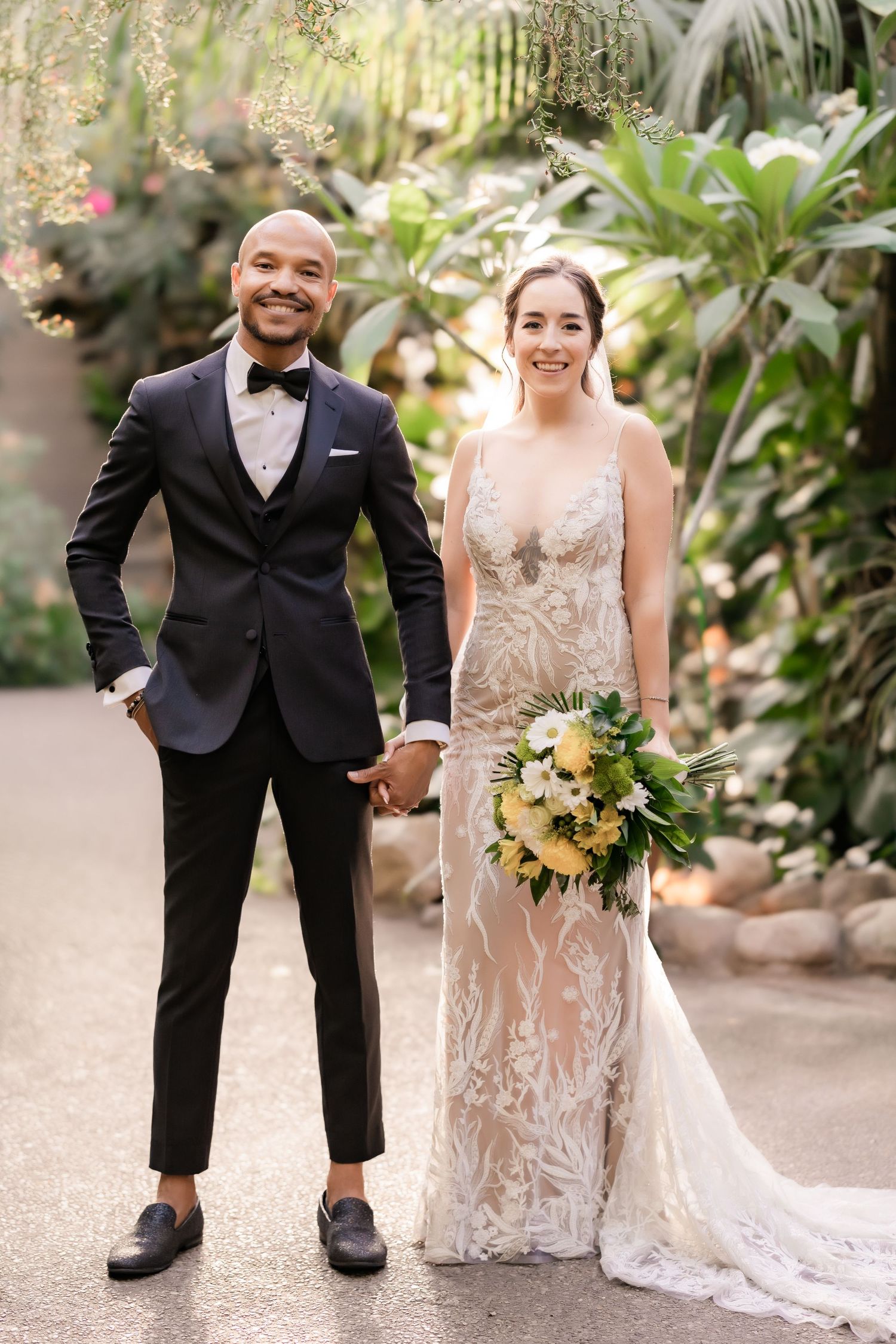 A bride and groom smile brightly while holding hands in a tropical garden setting.