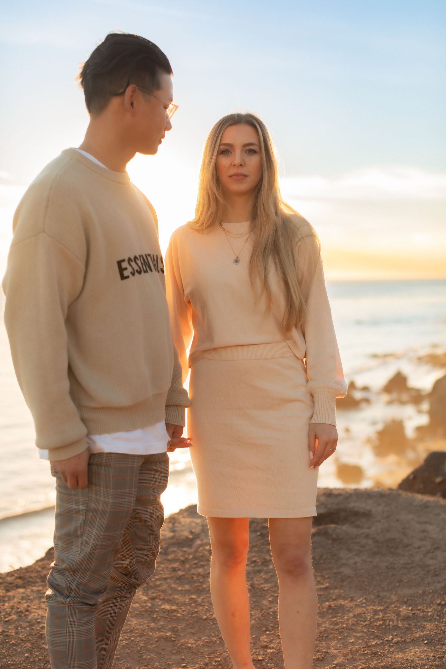 Couple in casual attire standing on coastal rocks at sunset.