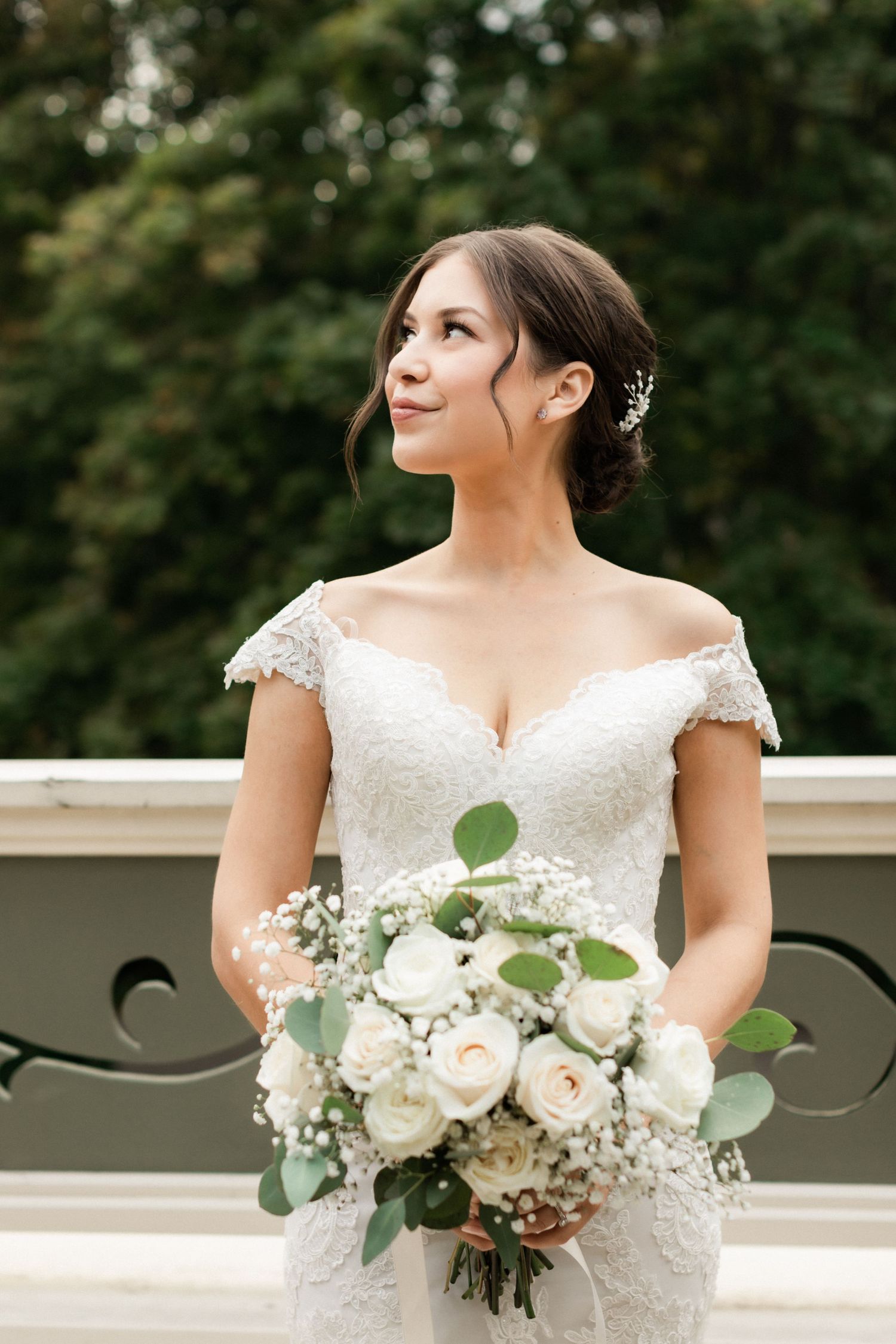 Elegant bride in off-shoulder lace gown holds white rose bouquet on outdoor balcony.