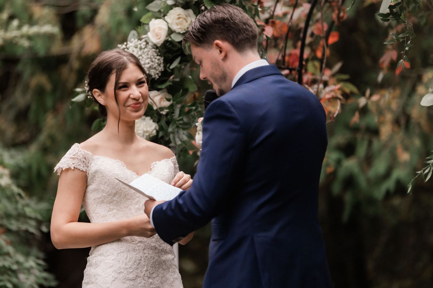 Beautiful outdoor wedding moment as couple exchanges rings surrounded by lush greenery and white floral decorations.